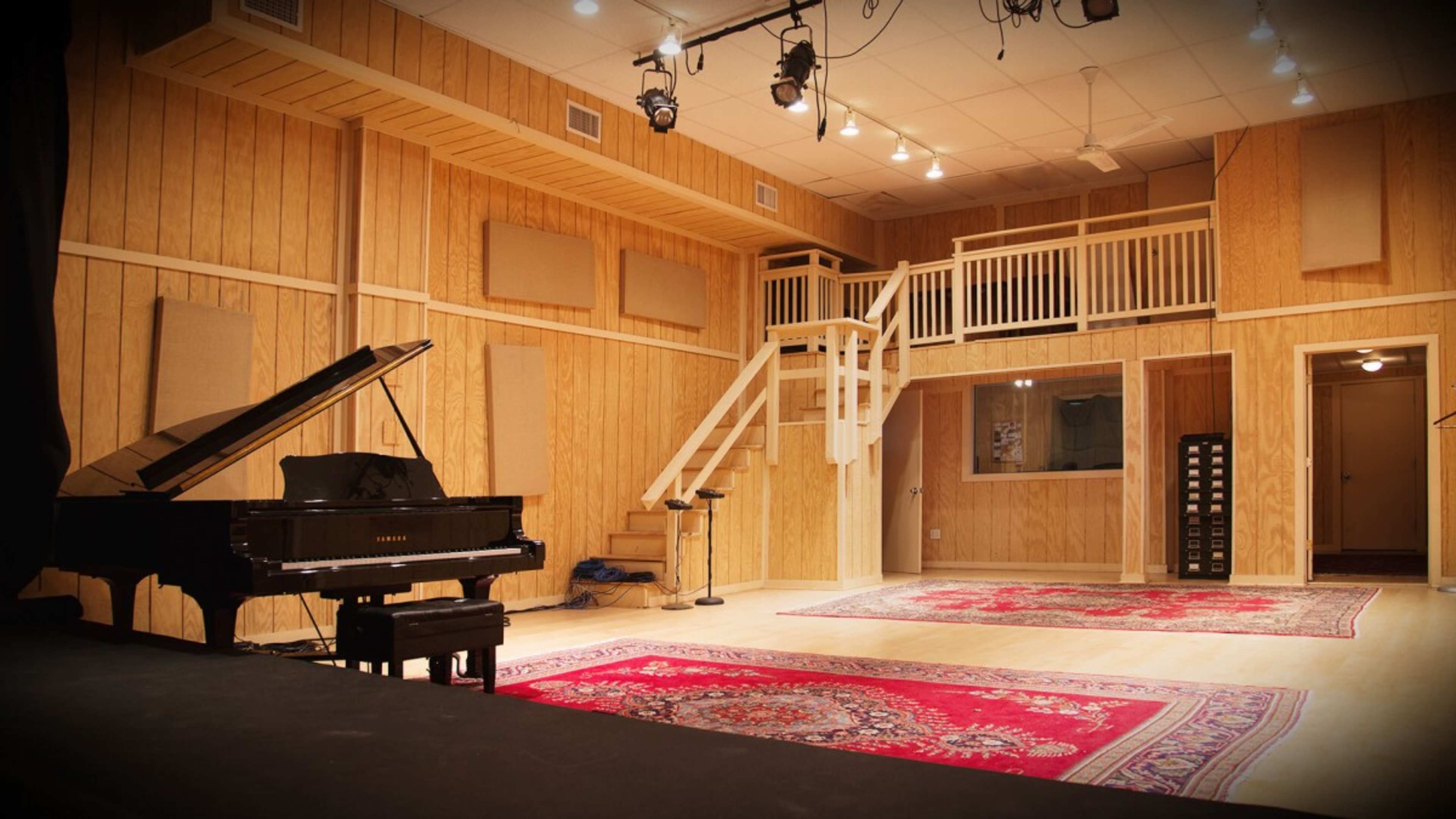 A music studio featuring a grand piano, wooden walls, a staircase, and carpeted areas with large rugs.