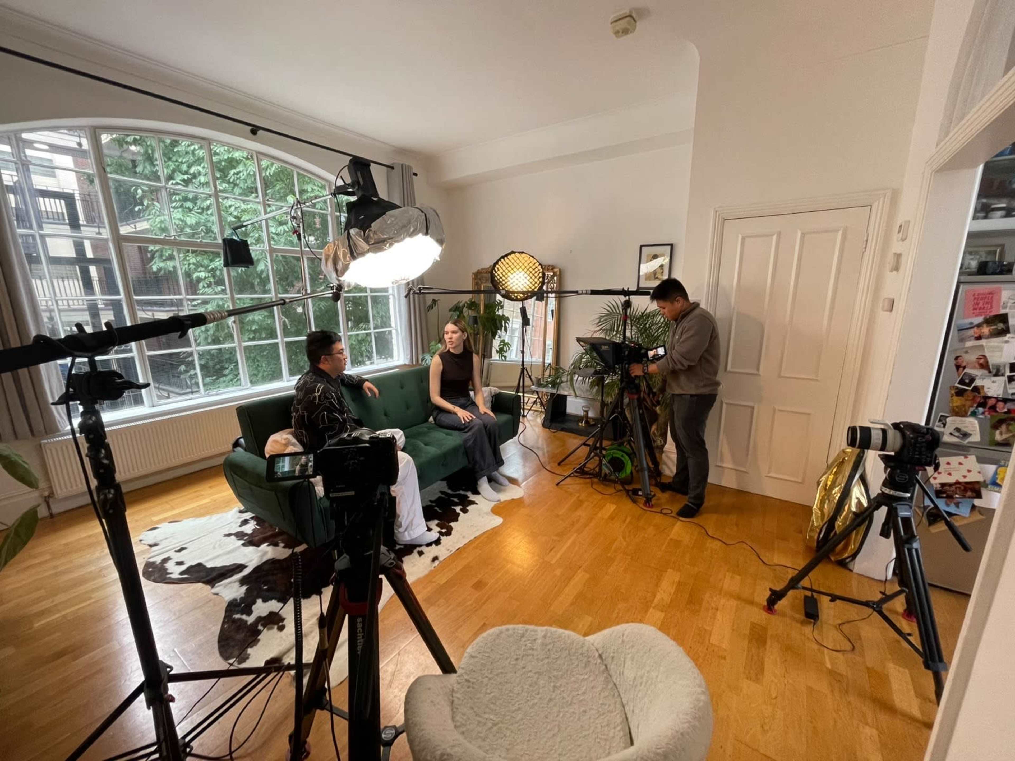 A modern living room is set up for a video shoot, featuring a green sofa, two people seated, and several cameras and lighting equipment arranged around the space.