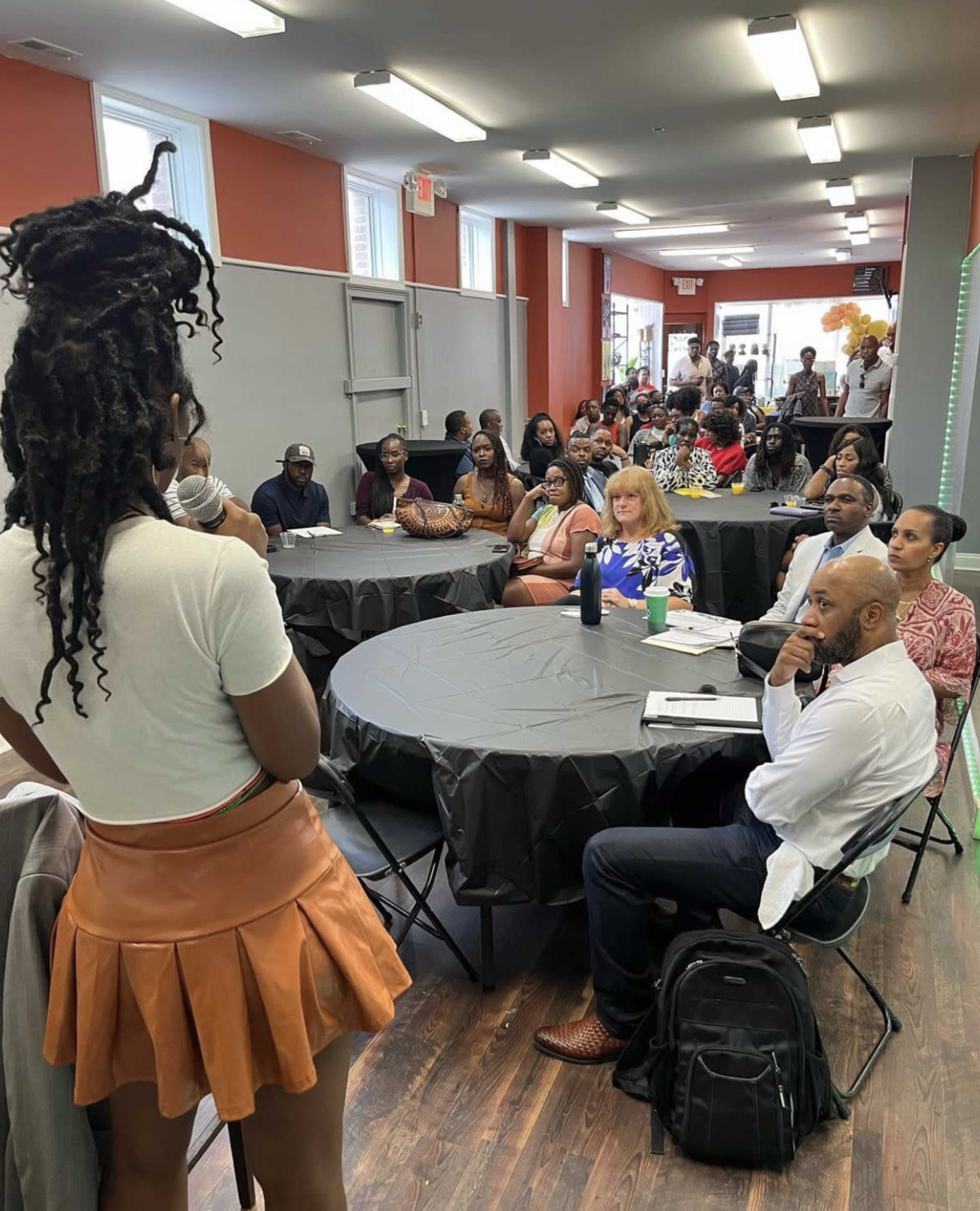 A speaker stands in front of a seated audience in a spacious, well-lit community room.