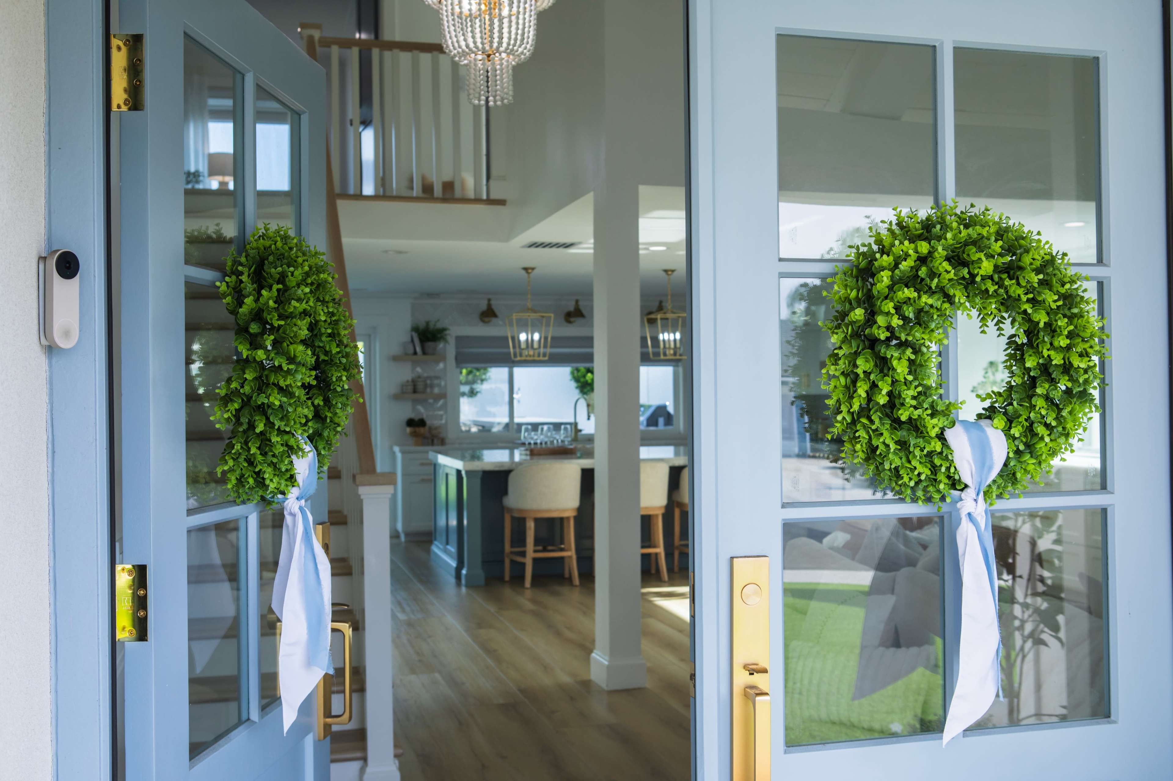 The image shows a pair of blue front doors with green wreaths adorned with blue ribbons, leading into a well-lit interior featuring a staircase and an open-concept living space.