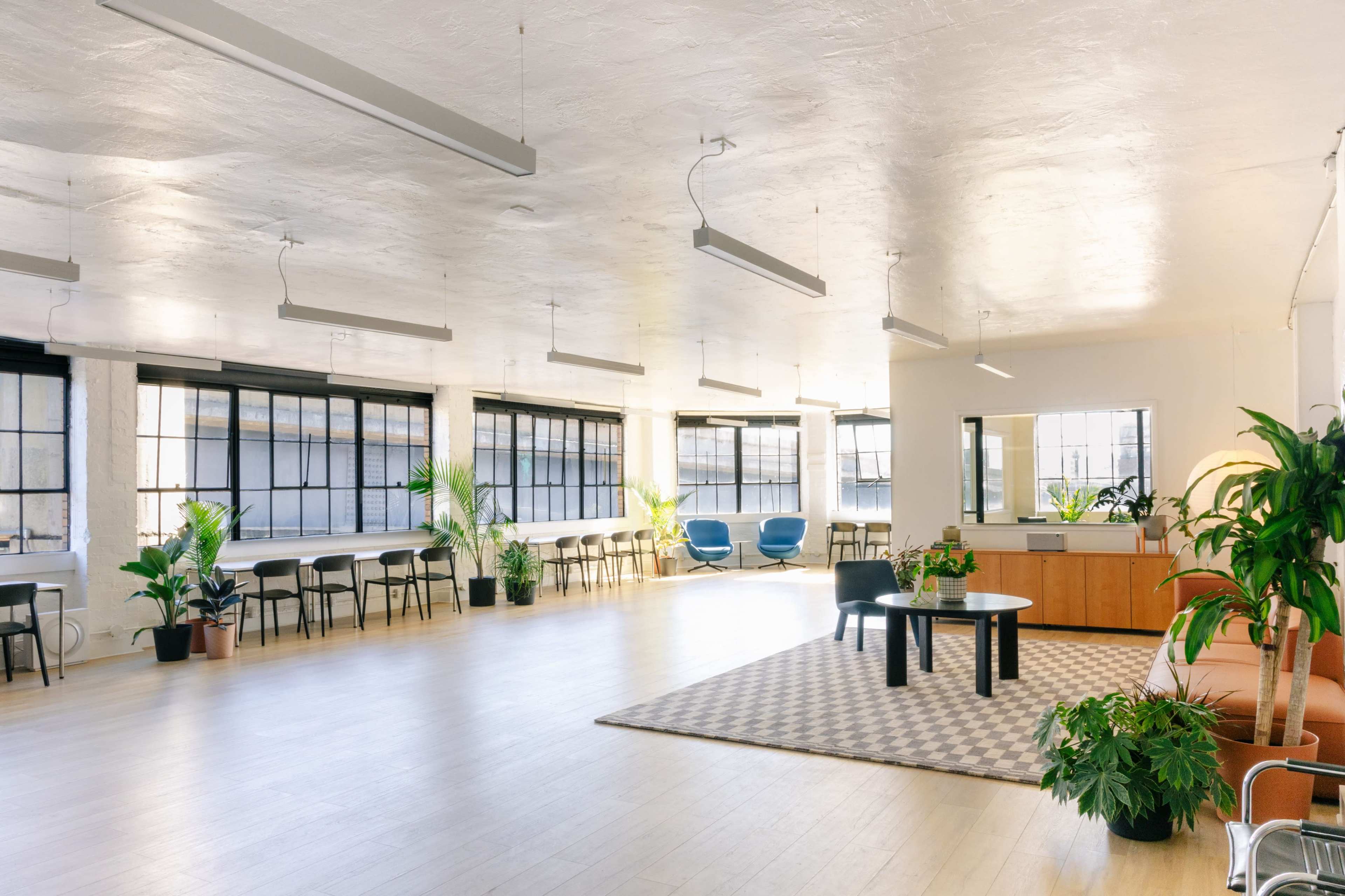 The image shows a spacious, well-lit modern office interior with large windows, plants, and various seating arrangements including tables and chairs.