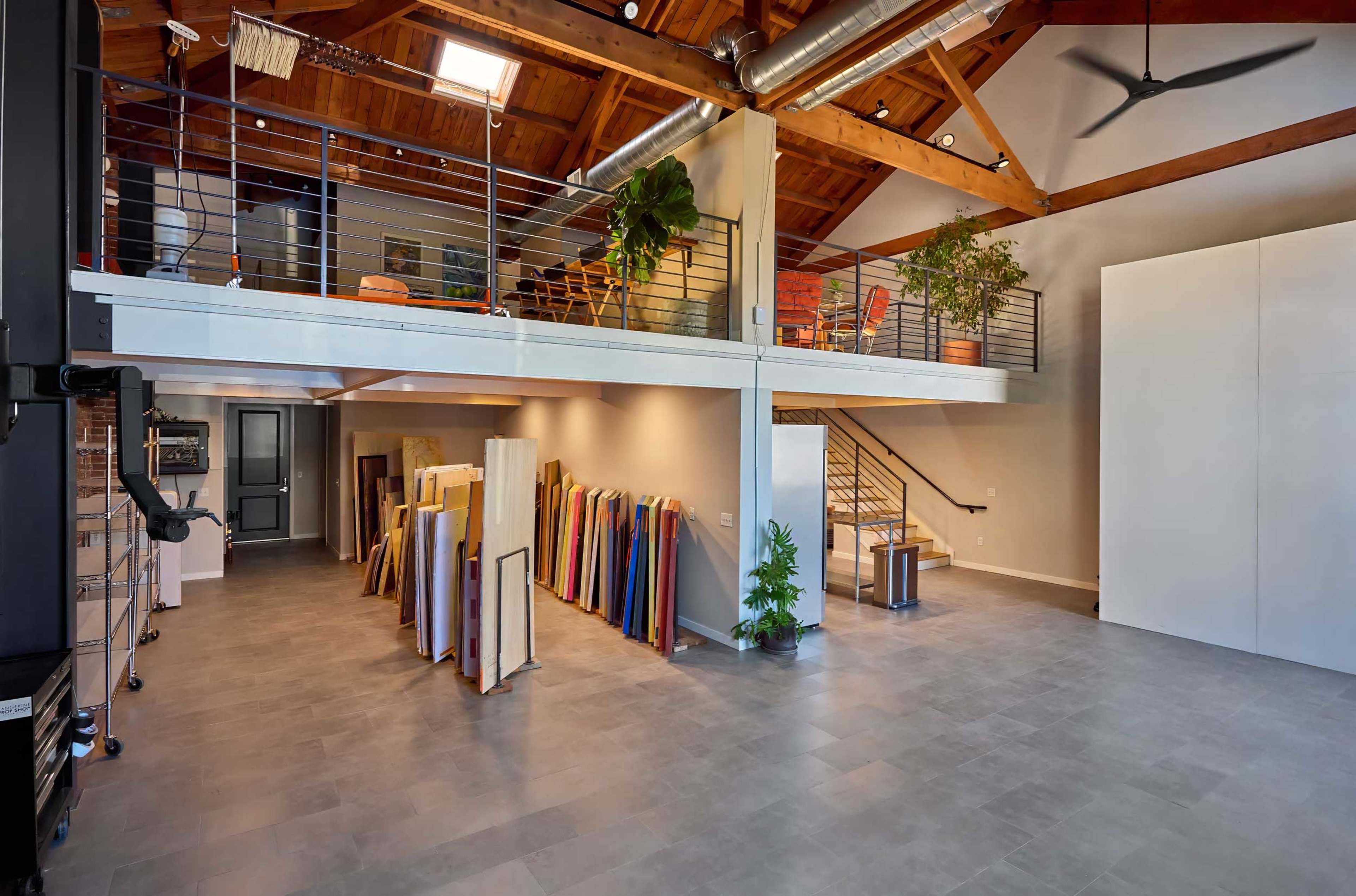 The image shows a spacious interior with a high wooden ceiling, featuring an upper loft area, a staircase, and stacks of colorful materials along one side.