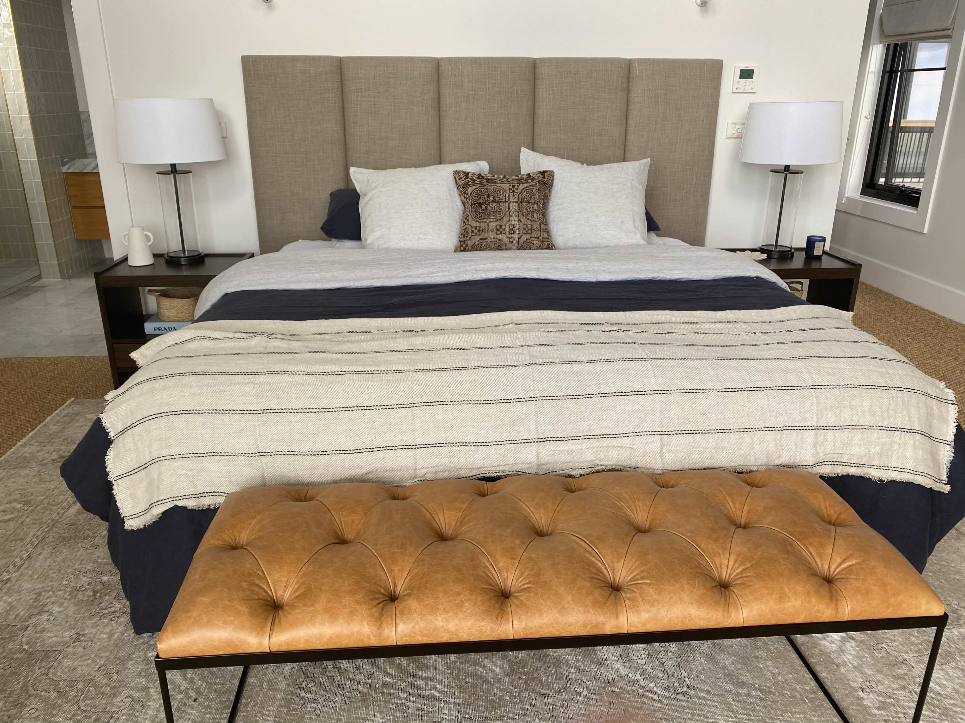 A modern bedroom featuring a large bed with a beige headboard, layered bedding, and a brown tufted bench at the foot of the bed.