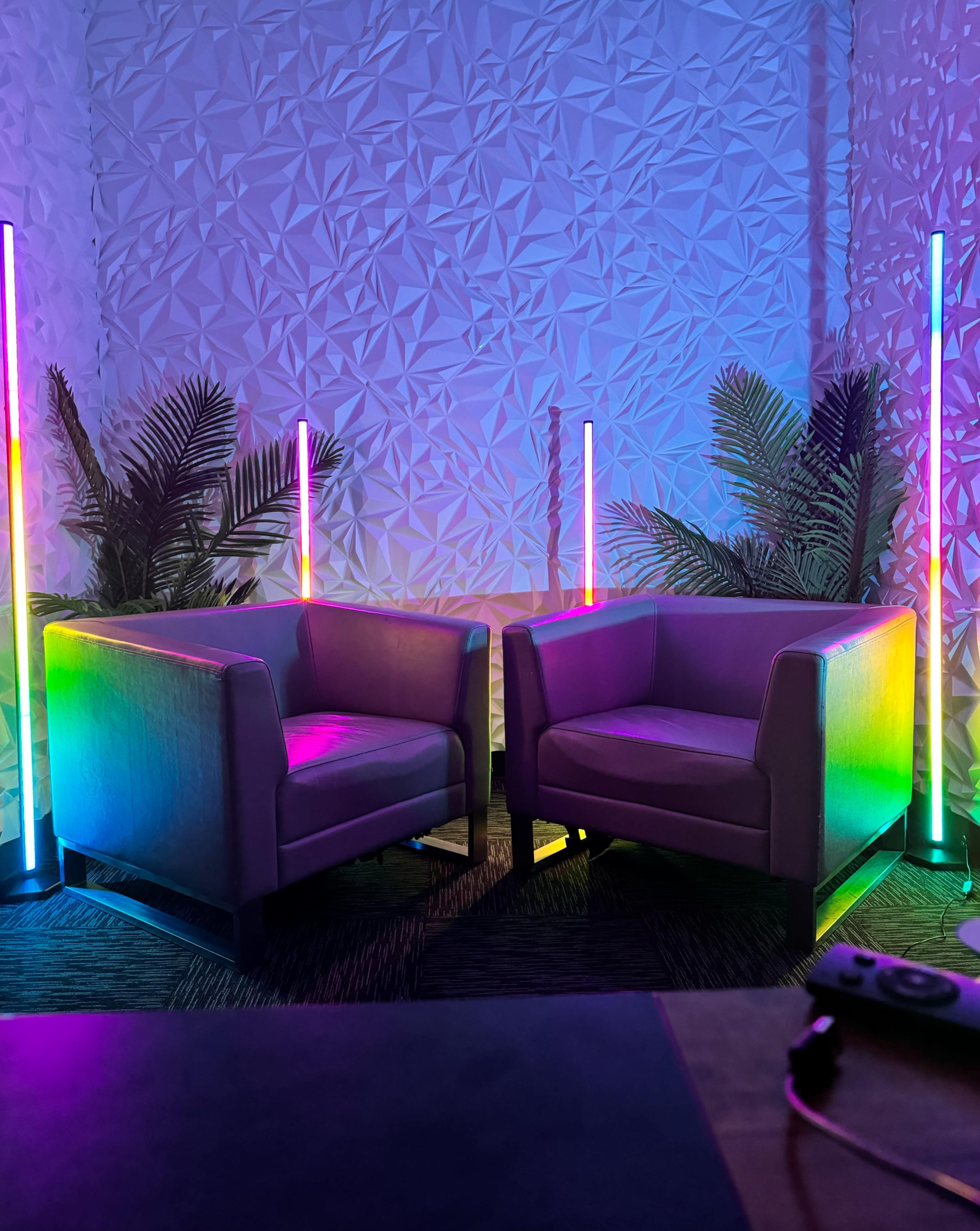 The image shows two modern chairs with colorful neon lights and decorative plants set against a textured wall.