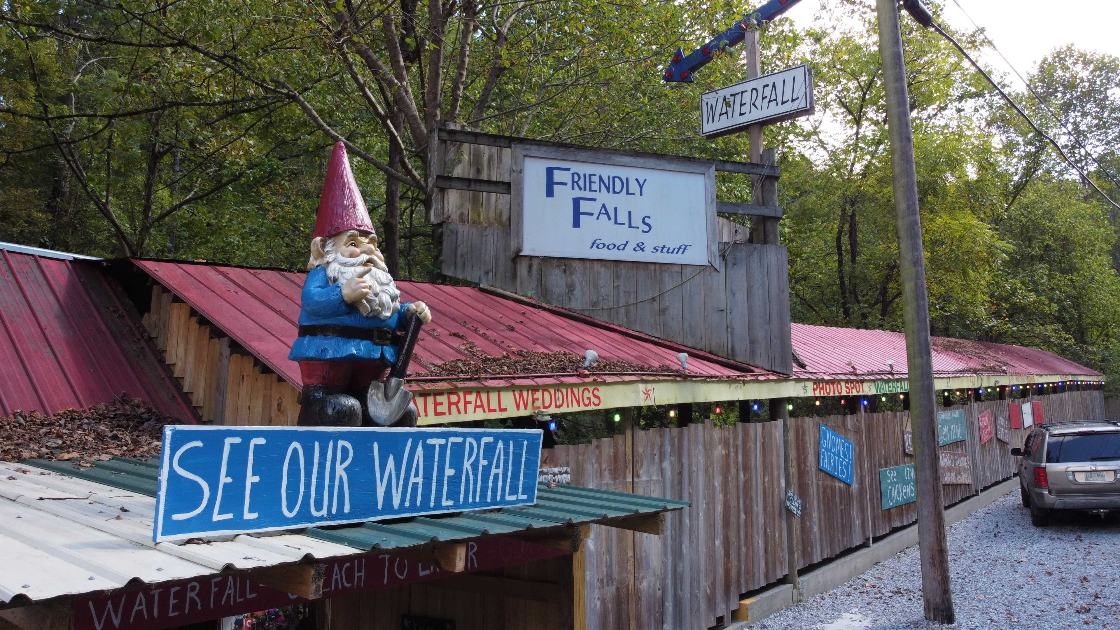 A rustic building featuring a gnome statue and signs advertising a waterfall and food services in a forested area.