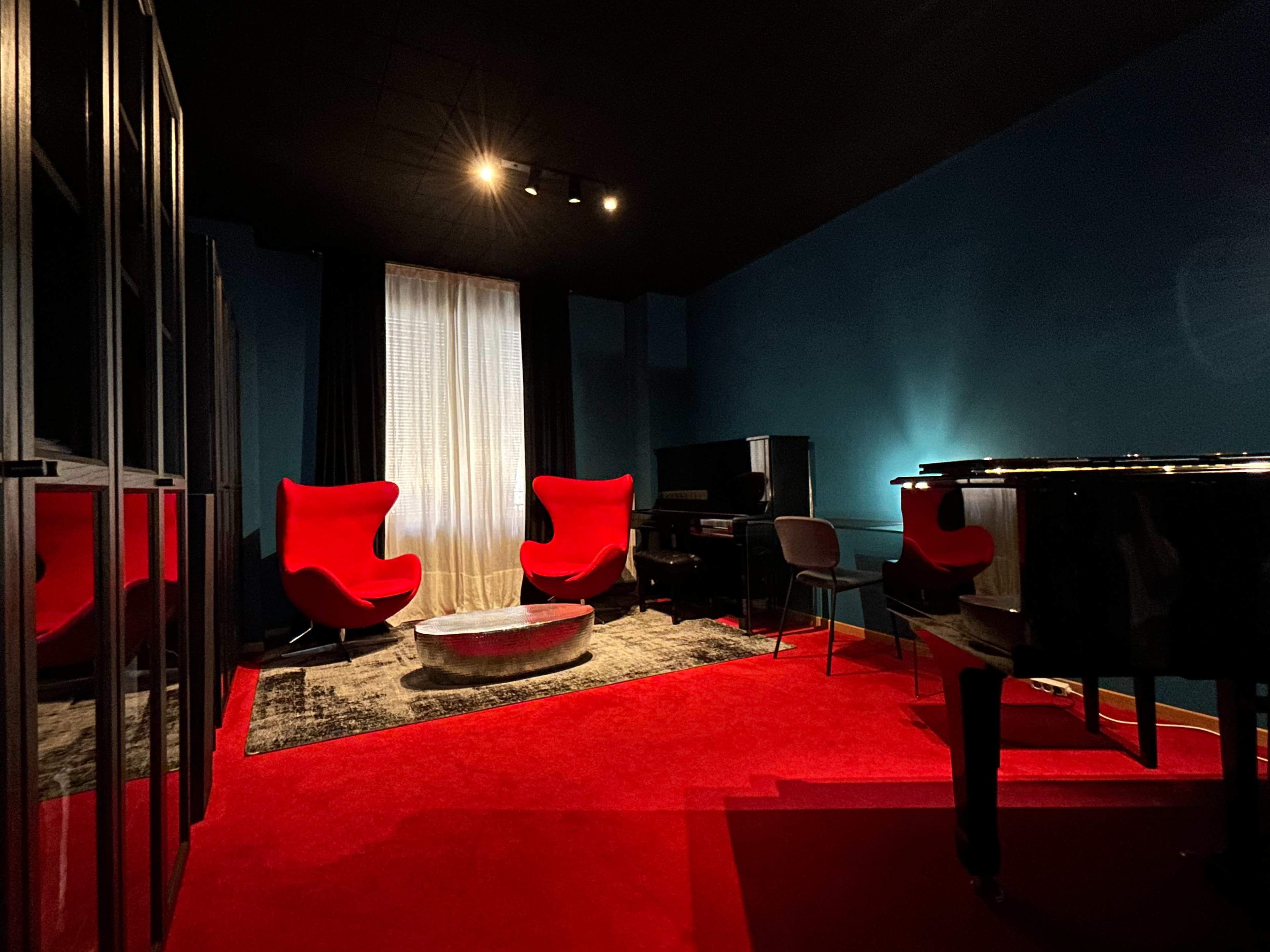 A music room features a black piano, two red accent chairs, a round table, and a large window with sheer curtains, all set against dark blue walls and a red carpet.