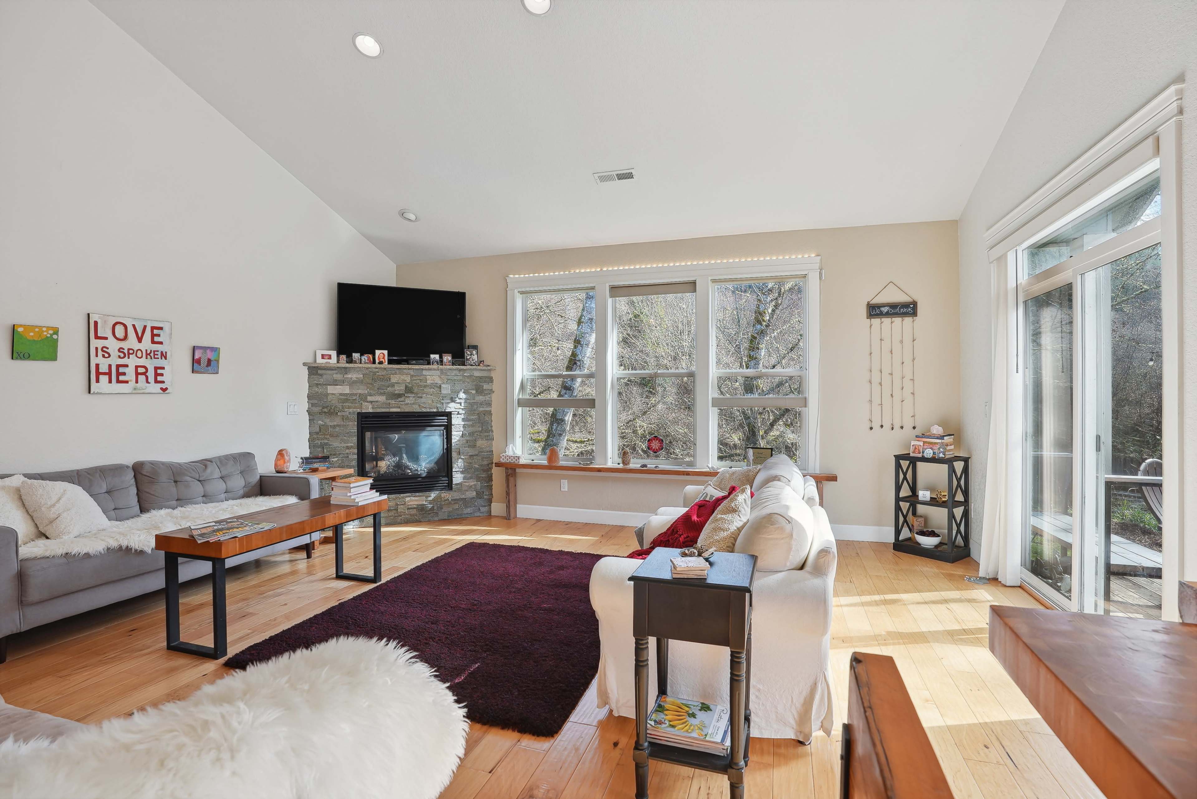 The living room features a stone fireplace, large windows, and a mix of furniture including a sofa, coffee tables, and a cozy rug.