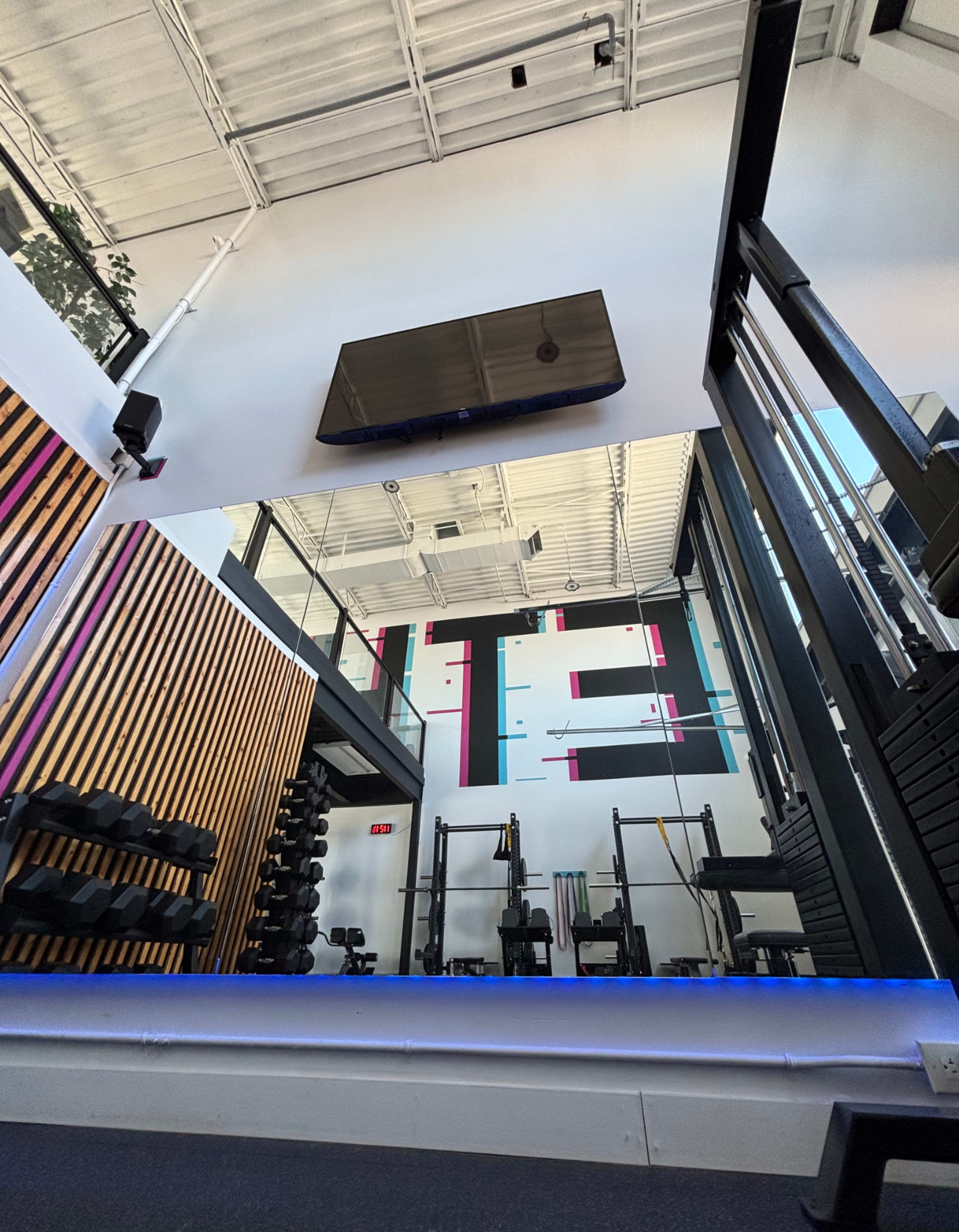 The image shows a modern gym interior with weight racks, exercise equipment, and a large wall-mounted television.