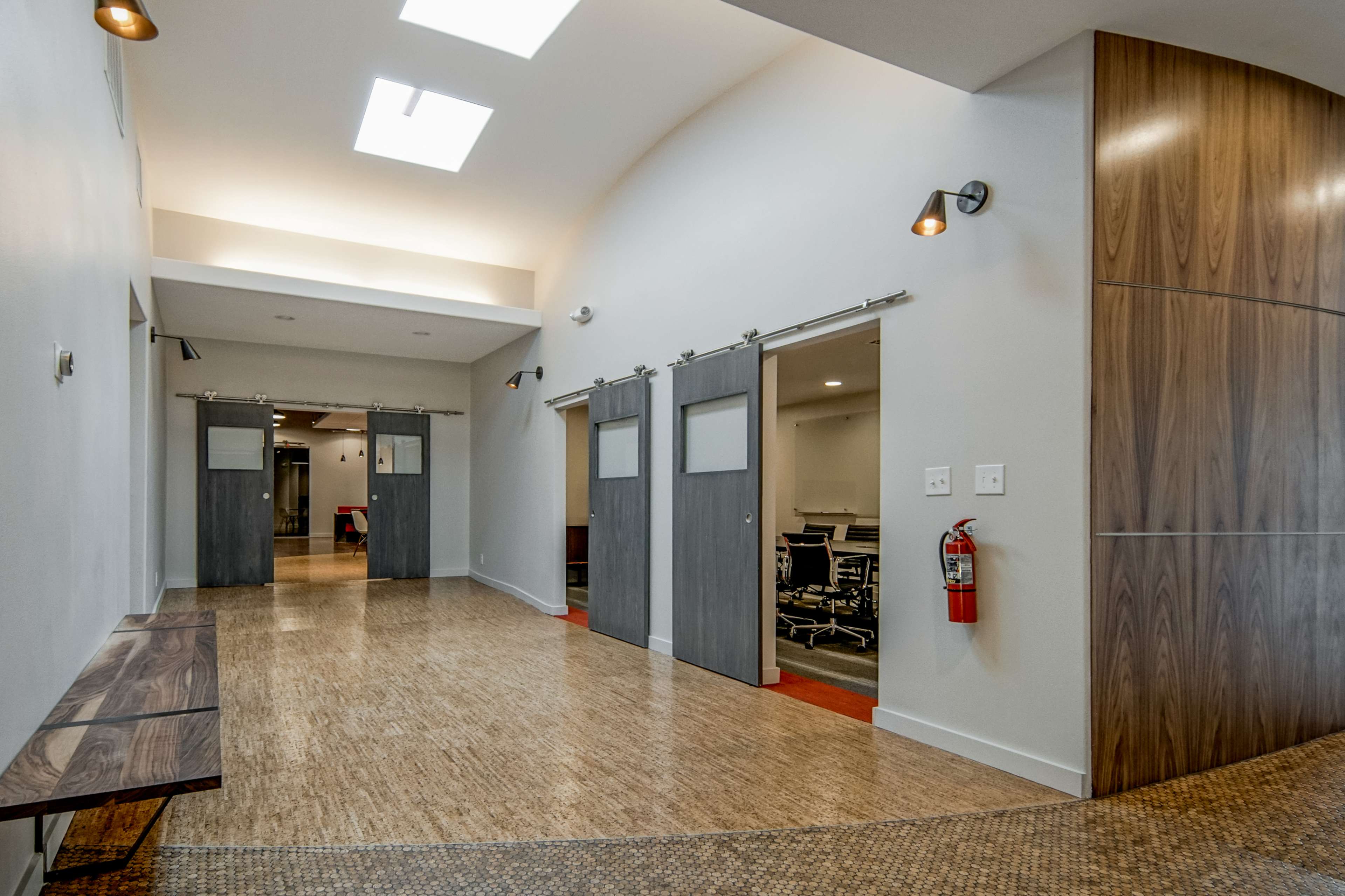 A modern hallway with sliding doors on either side, leading to office spaces, and a bench along one wall.