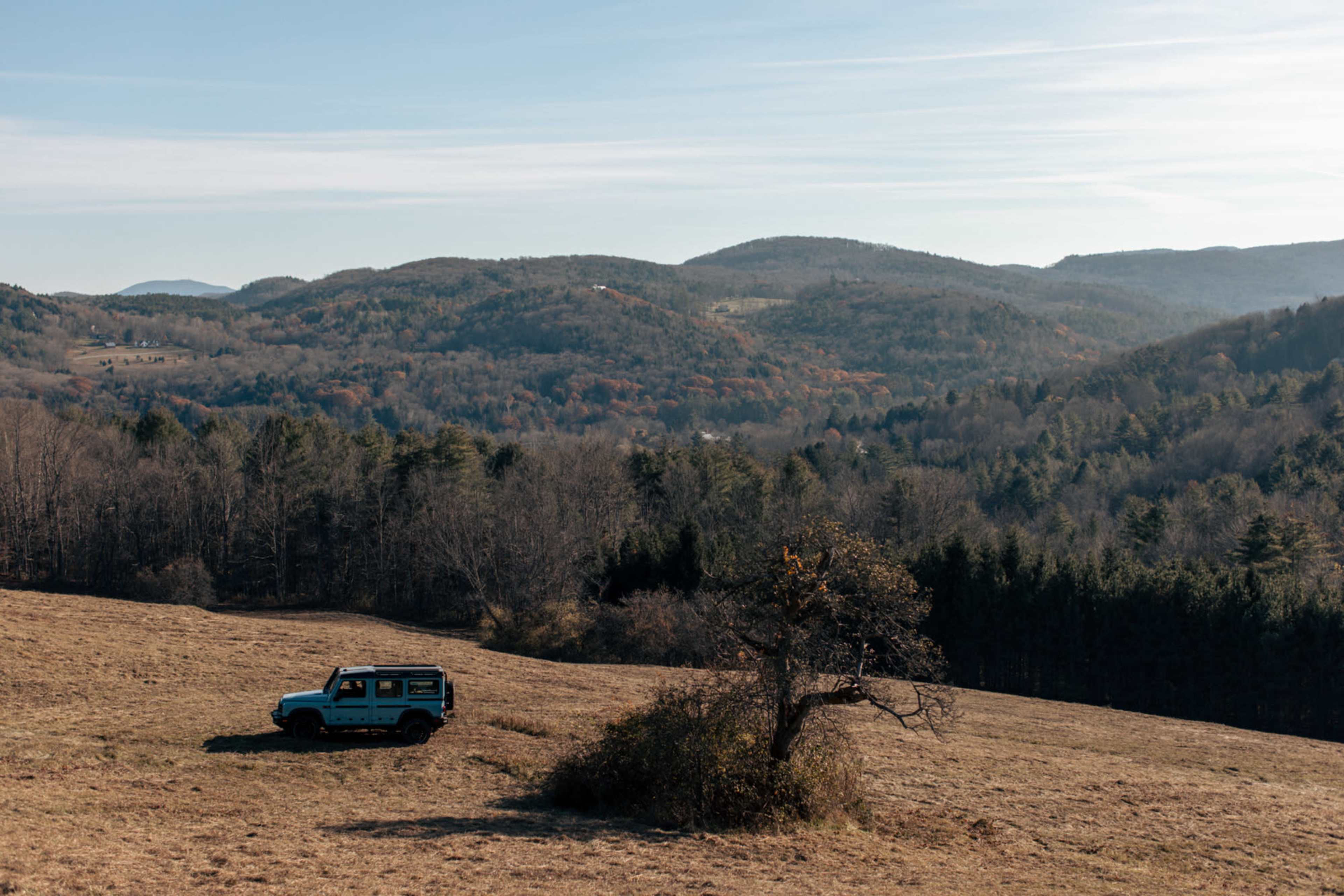 A parked SUV is situated in a grassy clearing surrounded by rolling hills and forests under a clear sky.
