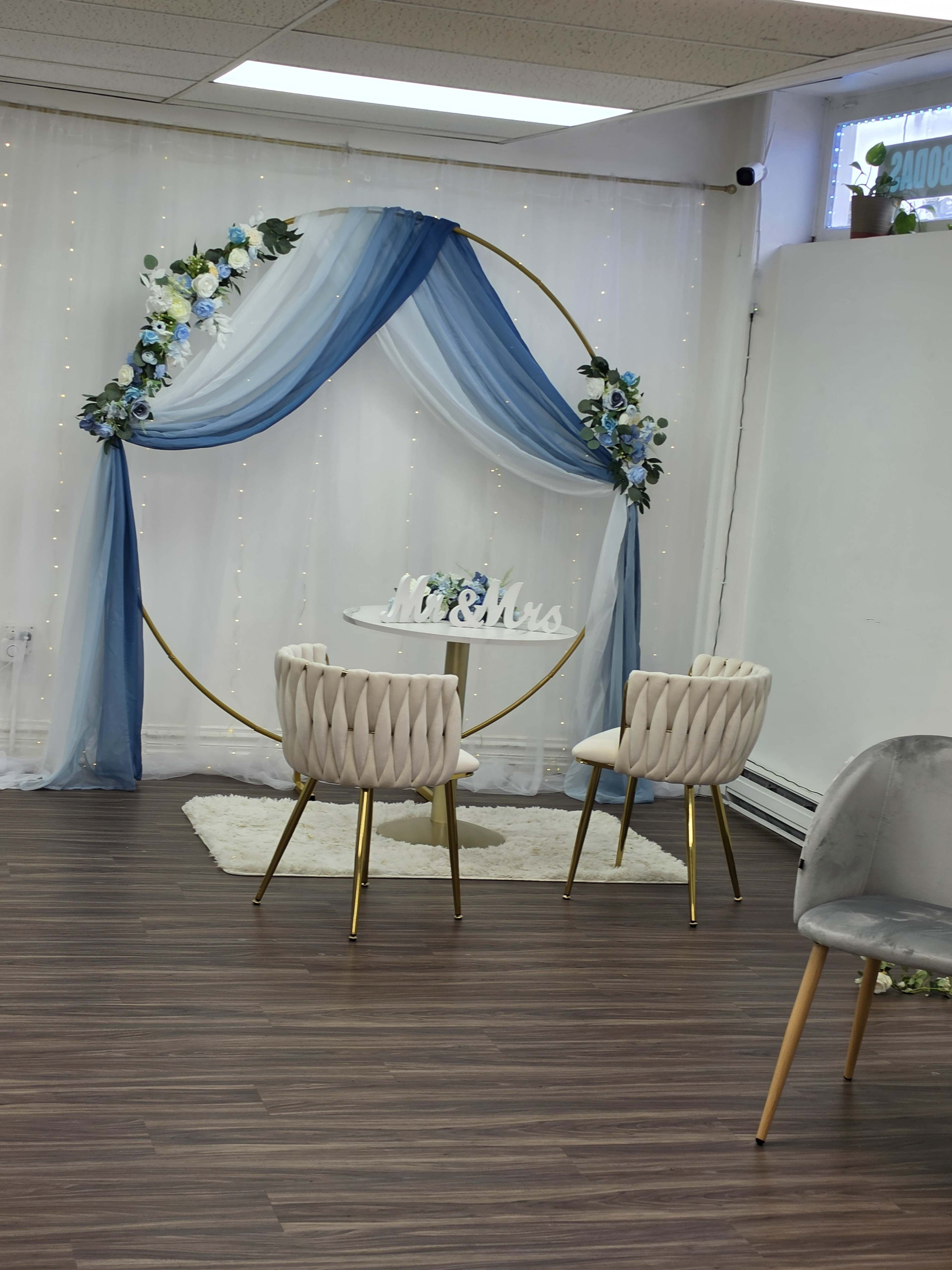 The image shows a wedding decor setup featuring a circular floral arch, a small table with a sign that reads "Mr. & Mrs.", and two modern chairs arranged on a white rug.