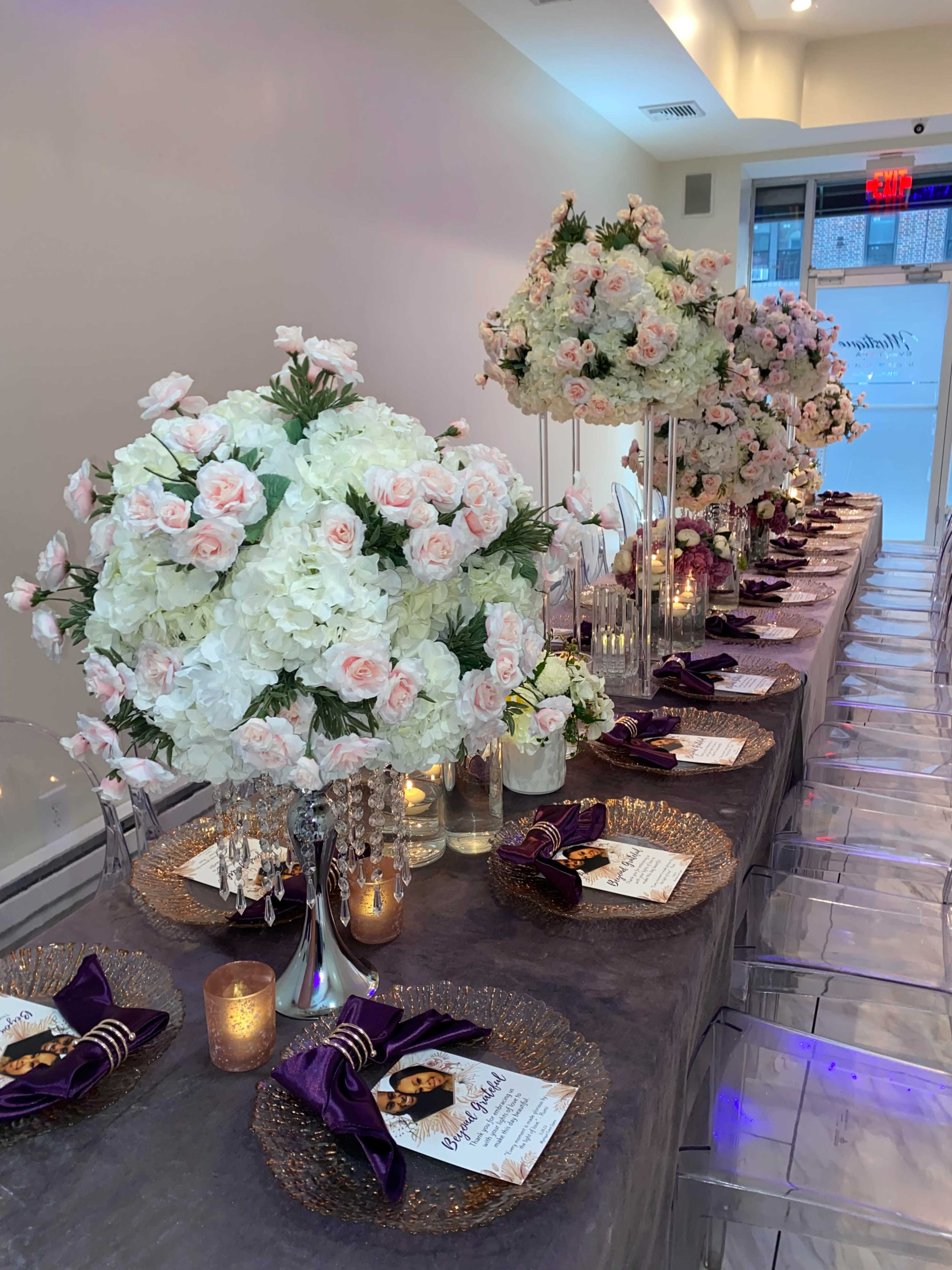 A long dining table is elegantly set with floral centerpieces, golden charger plates, and purple napkins.