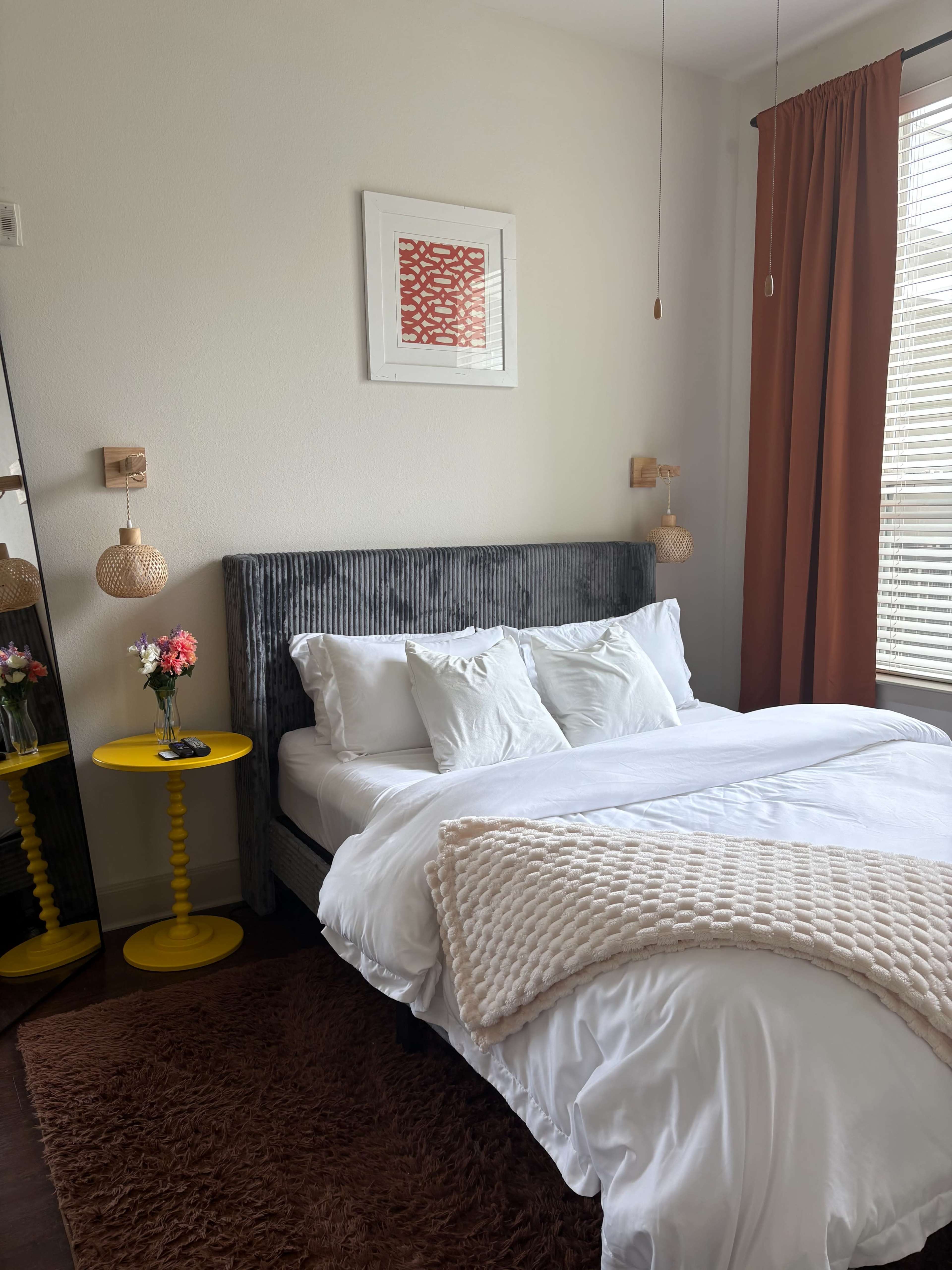 The image shows a neatly arranged bedroom with a gray upholstered bed, white bedding, a yellow side table with a flower vase, and a window with sheer curtains.