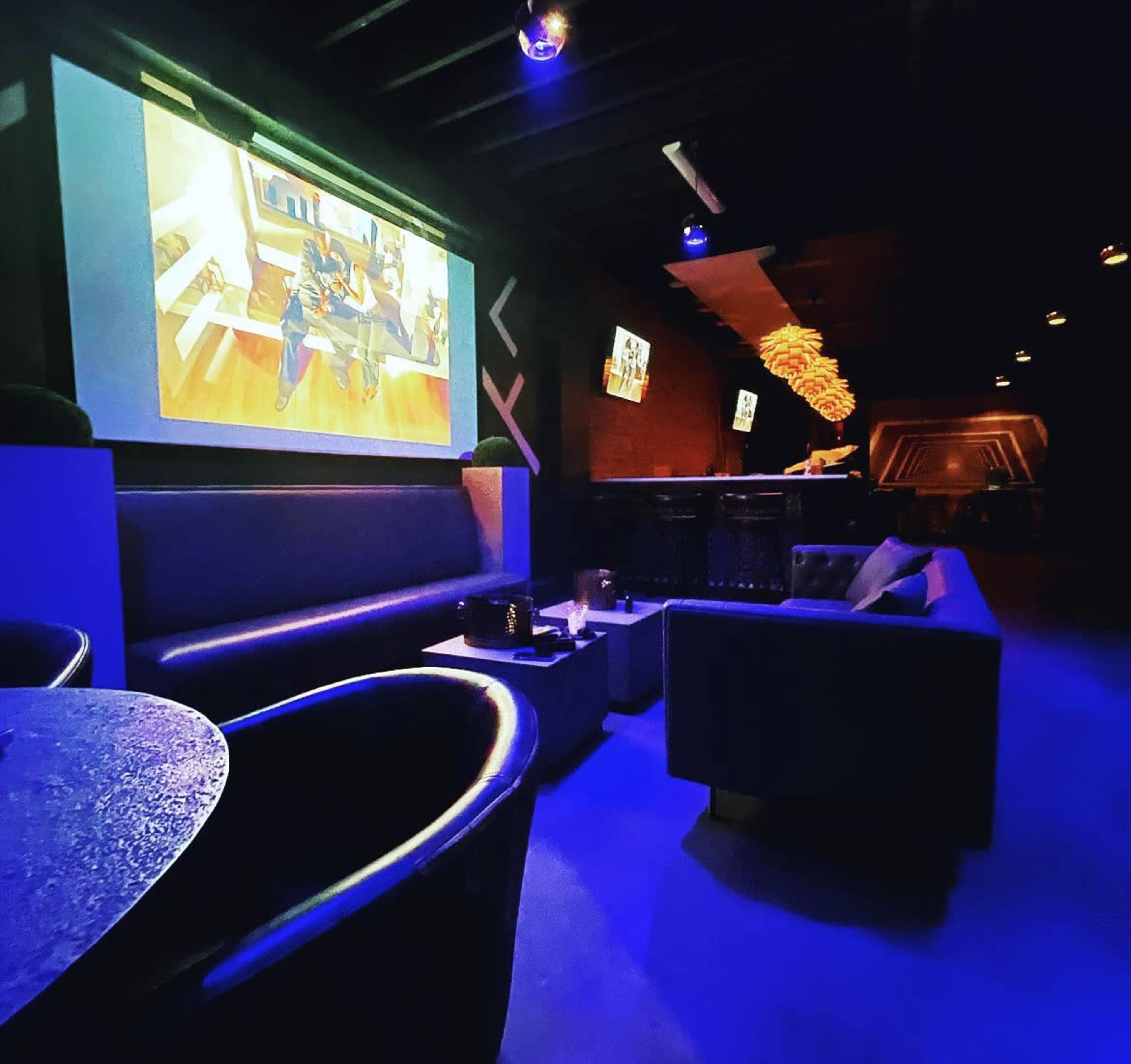 A dimly lit lounge area with dark furniture and a large screen displaying an animated scene on the wall.