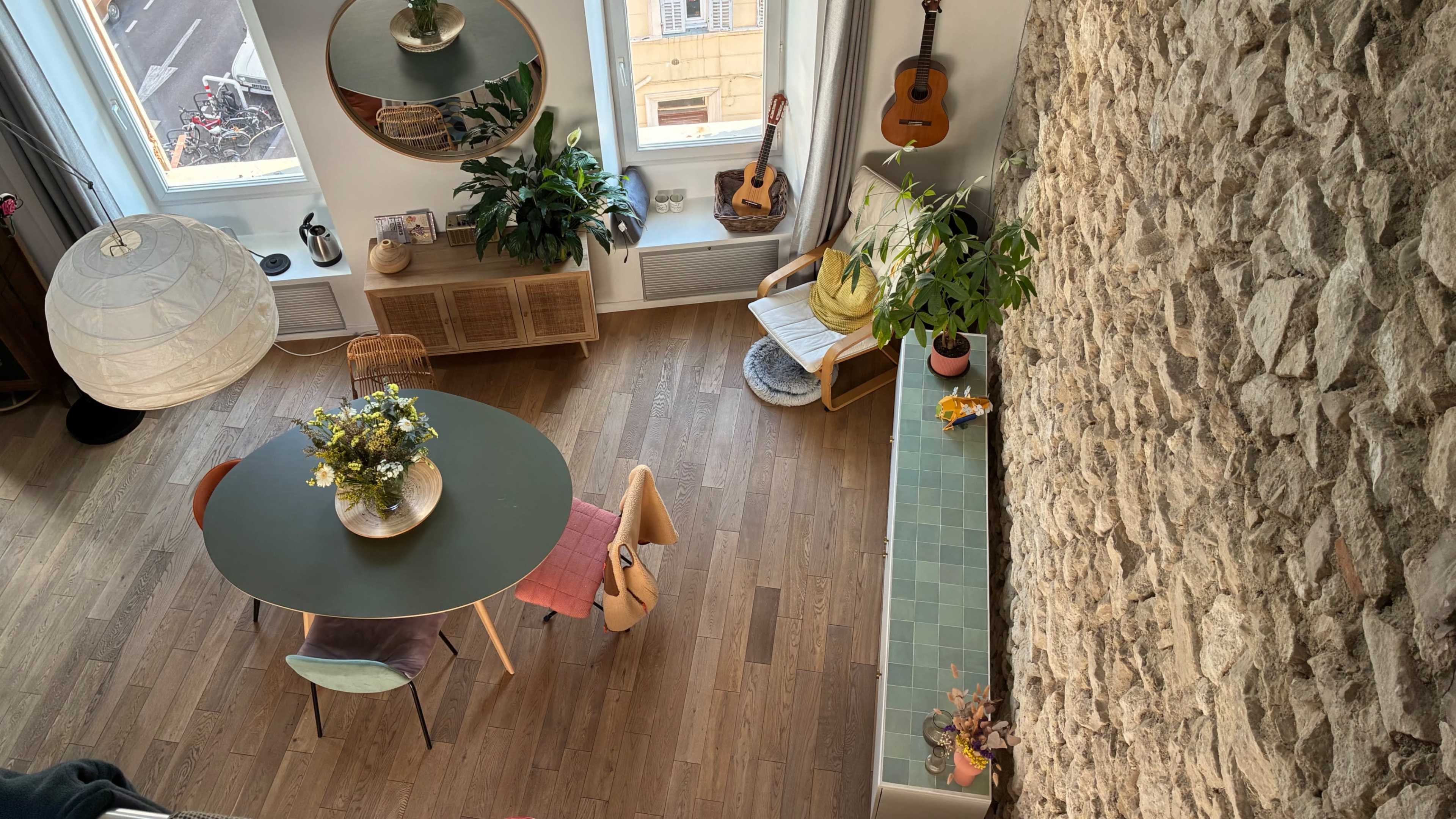 The image showcases an overhead view of a modern living space featuring a round table surrounded by colorful chairs, large windows, plants, and a stone wall.