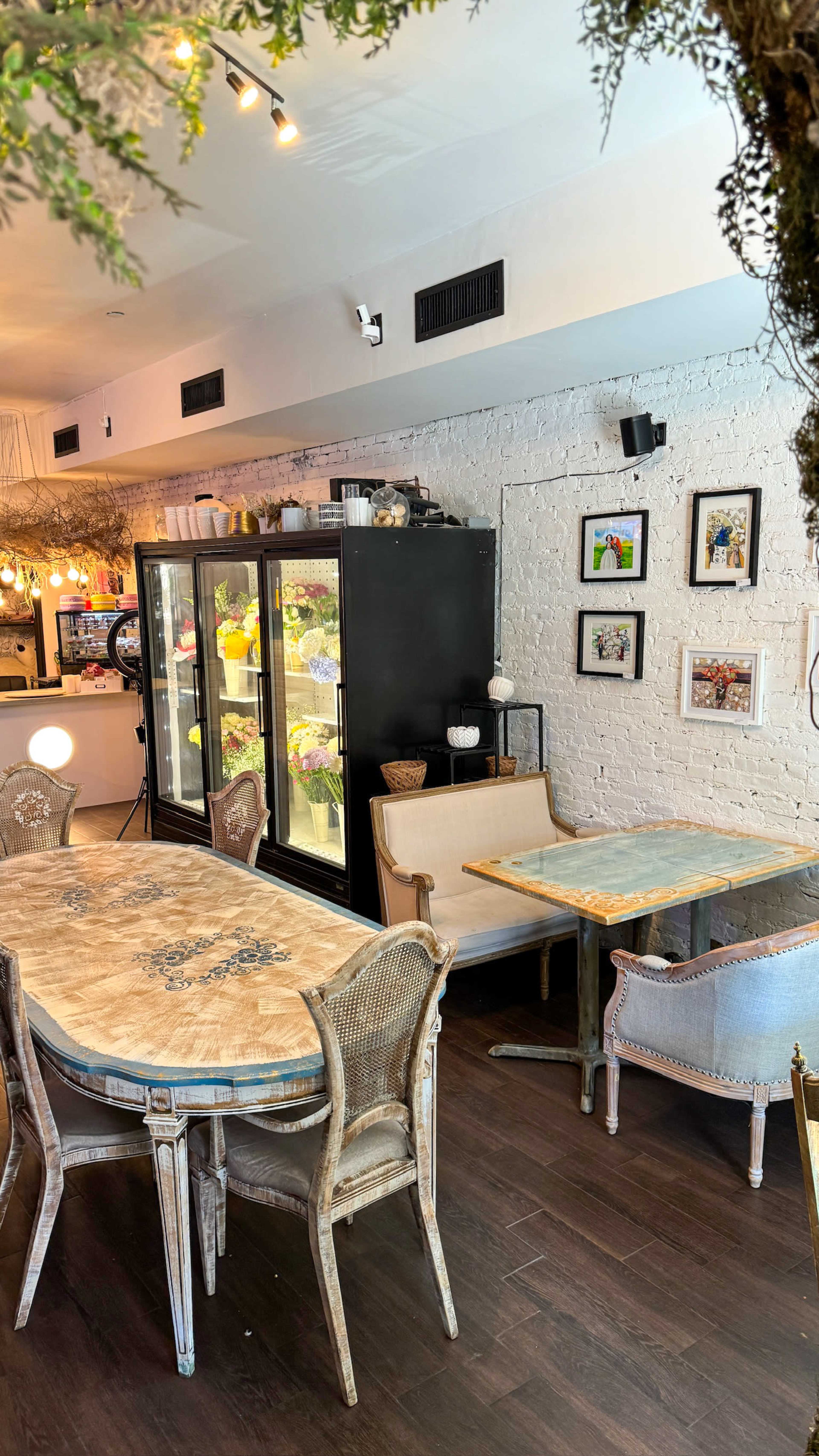 A café interior featuring wooden tables and chairs, a display refrigerator, and decorative framed pictures on the wall.