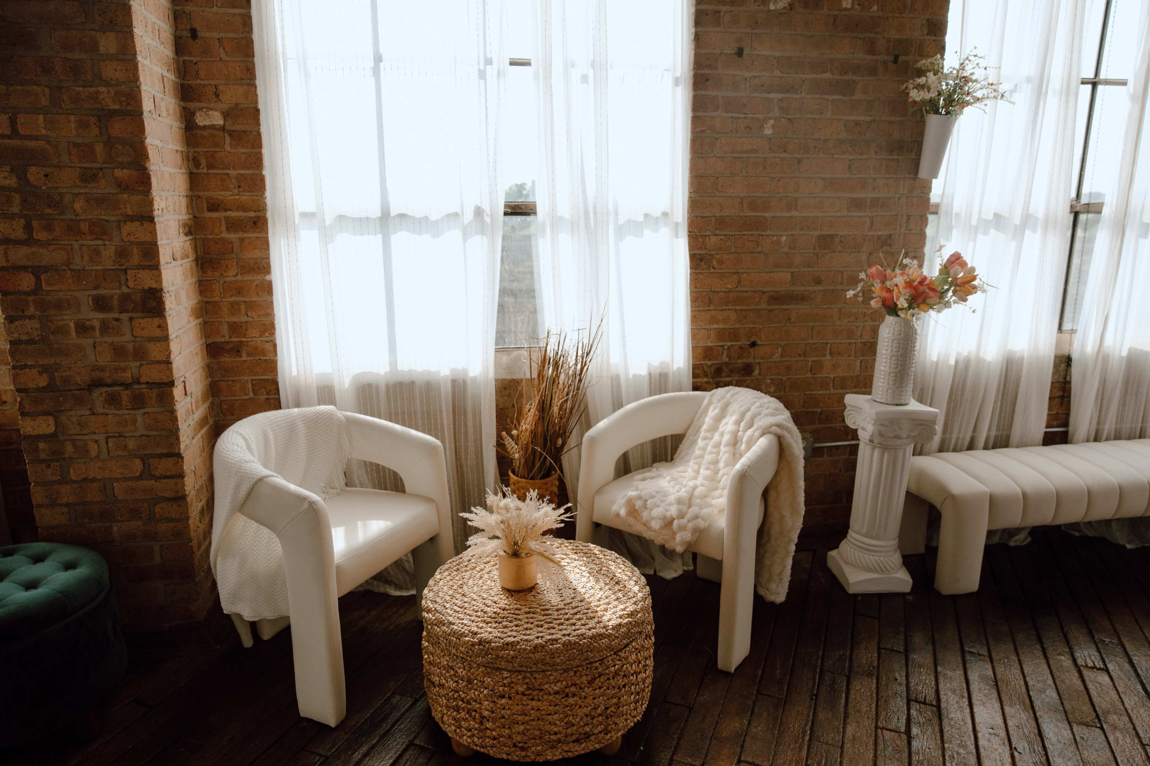 A cozy seating area features two white chairs and a woven coffee table surrounded by brick walls and sheer curtains.