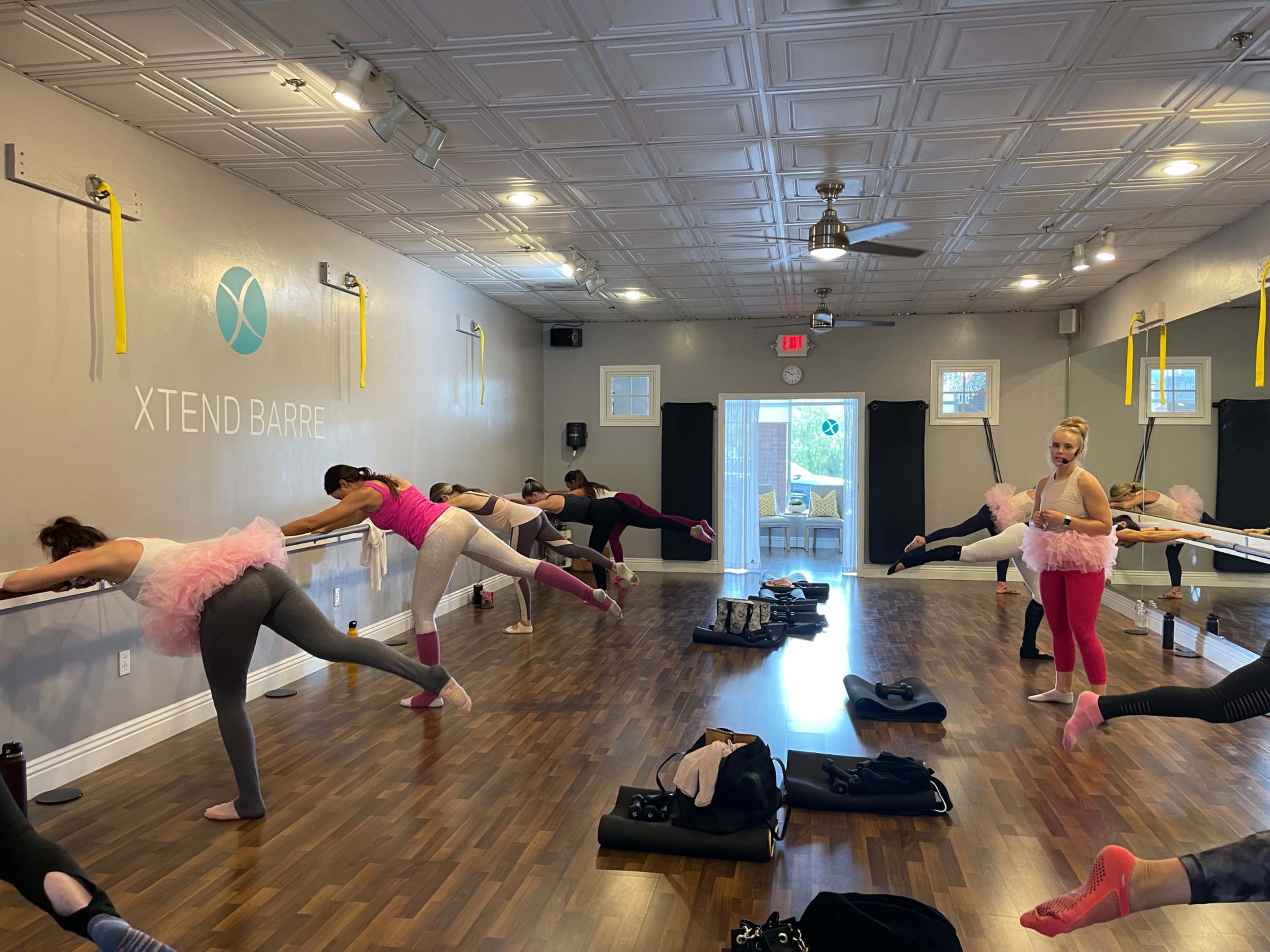 A group of women in a fitness studio participates in a barre class, using wall-mounted bars for support while performing leg exercises.