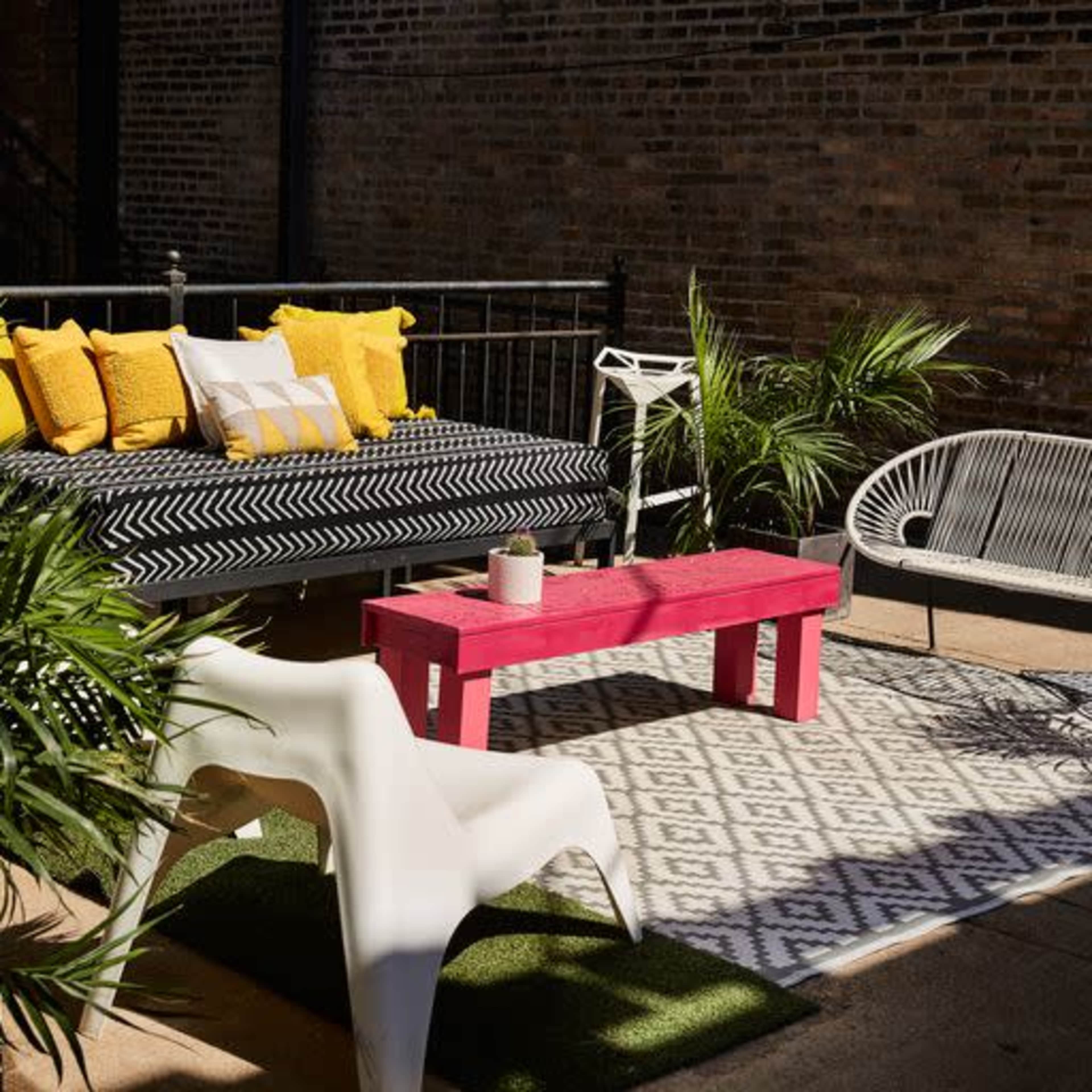 The image shows a modern outdoor seating area featuring a patterned rug, a pink coffee table, and various plants.