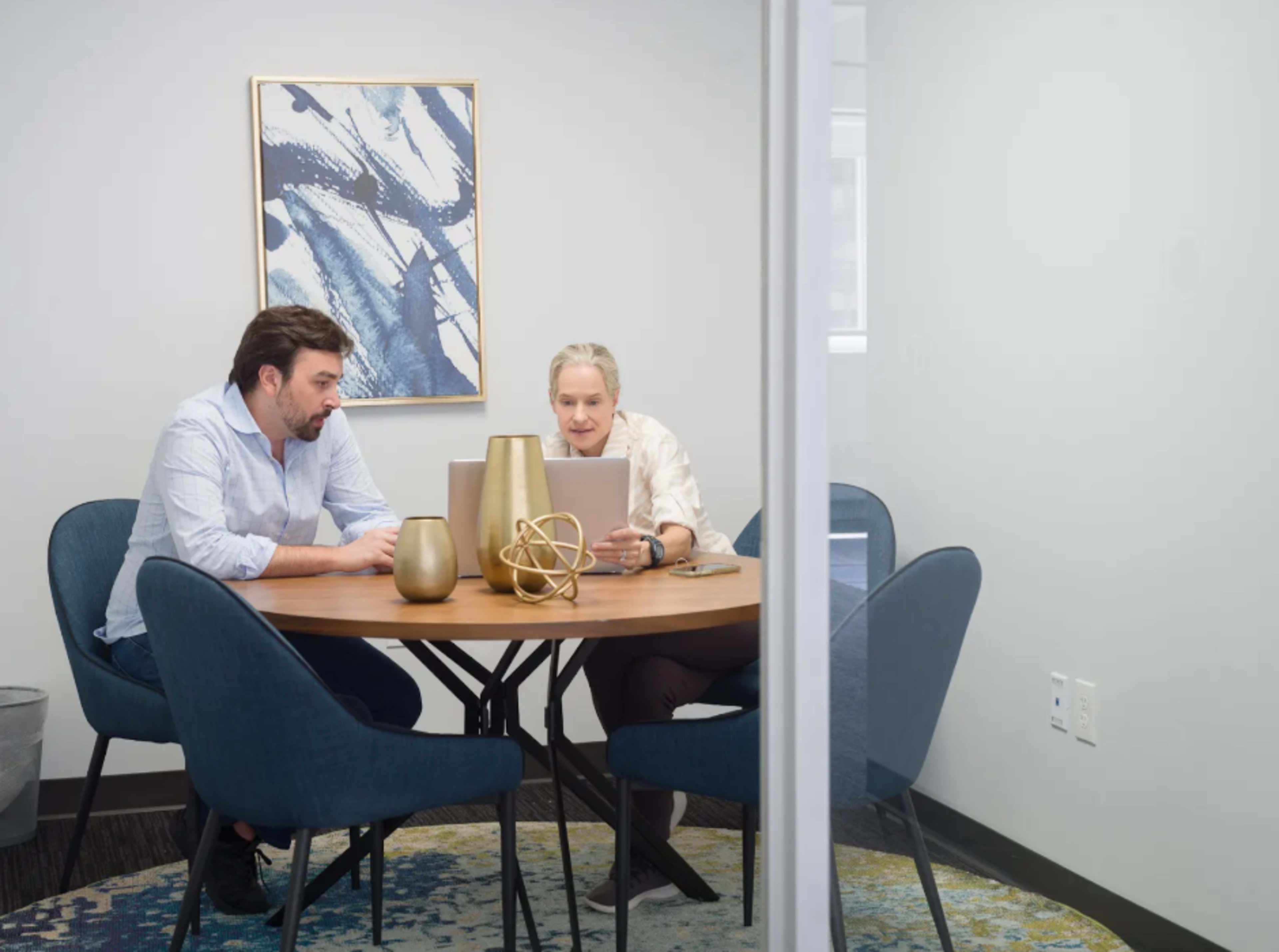 Two people are sitting at a round wooden table working on laptops in a small conference room.
