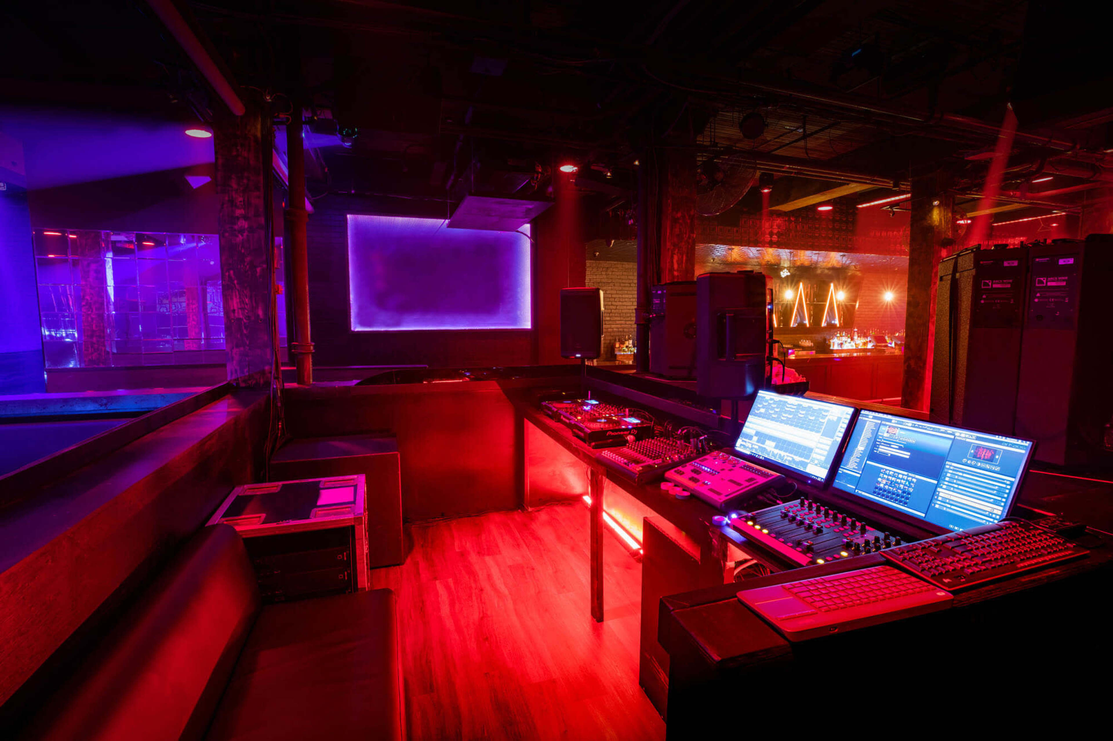 The image shows a dimly lit DJ booth in a nightclub, featuring multiple digital equipment and colorful lighting effects.