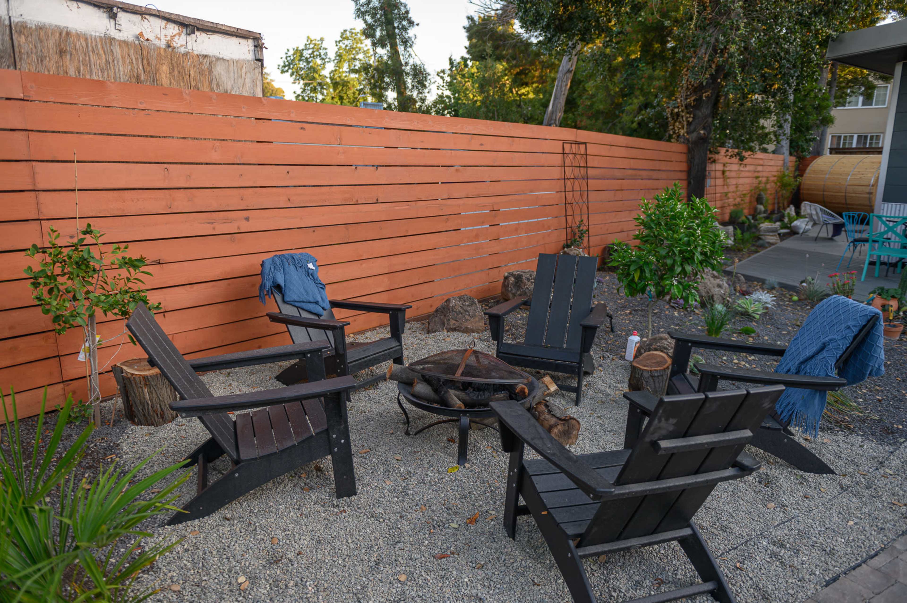A cozy outdoor seating area features four black Adirondack chairs arranged around a fire pit on a gravel surface, with a wooden fence in the background.