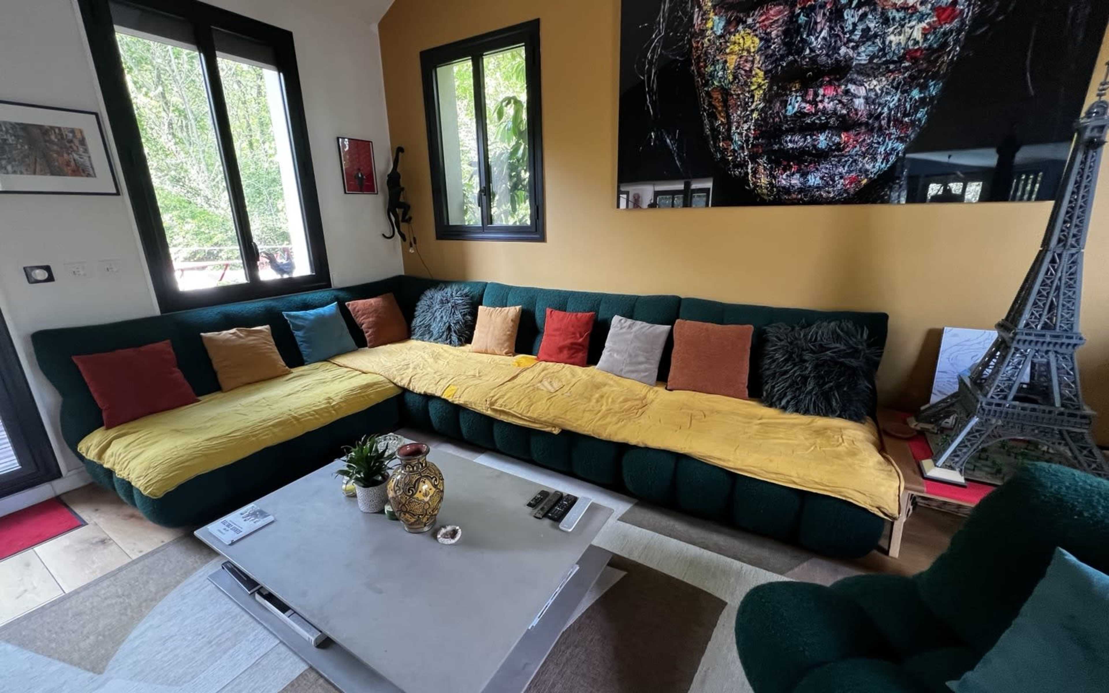 The image shows a spacious living room featuring a large green sectional sofa adorned with colorful cushions, a coffee table in the center, and a vibrant wall art piece.