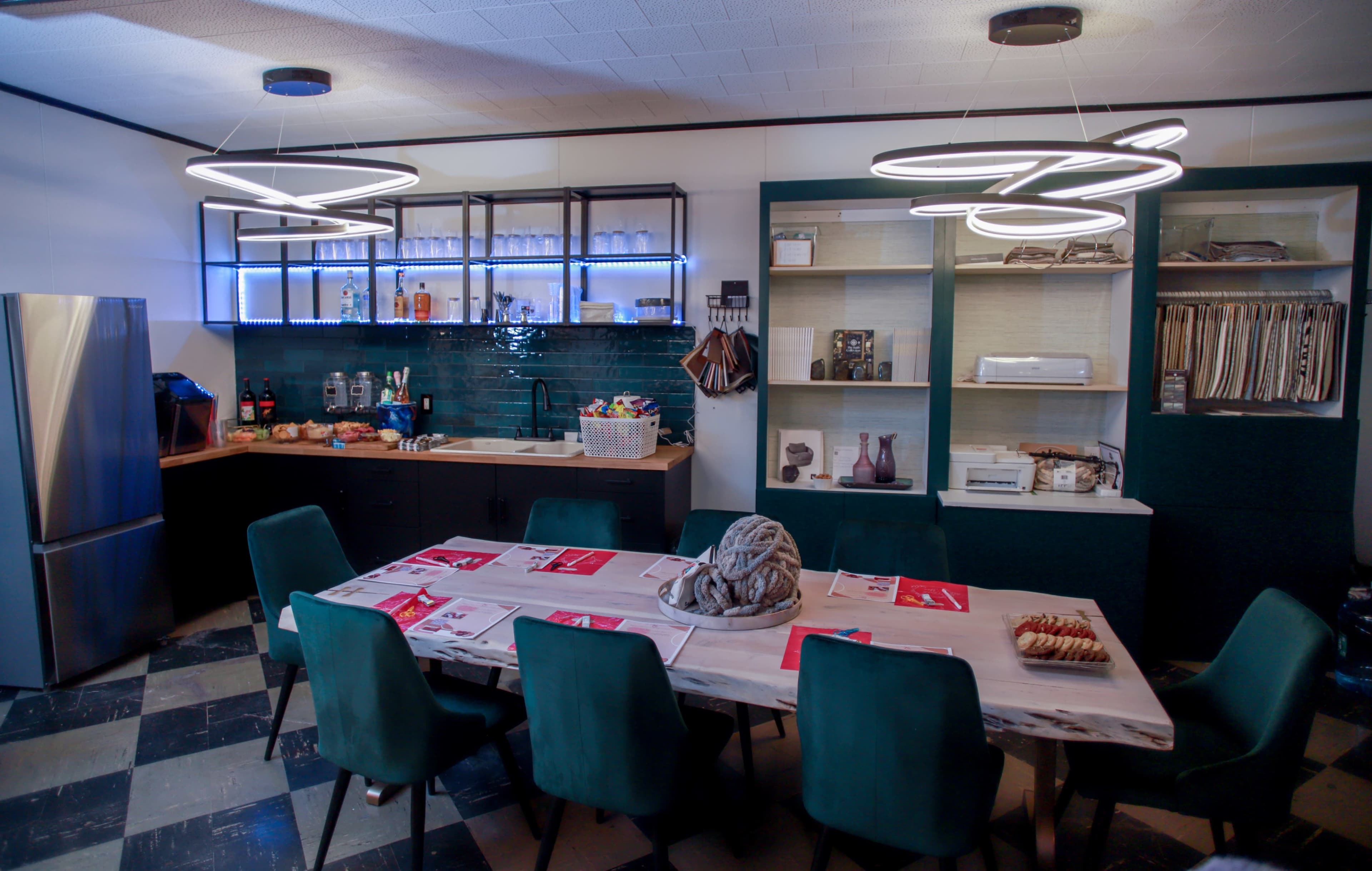A modern kitchen features a large dining table surrounded by green chairs, with a refrigerator, dark cabinetry, and illuminated shelves displaying various kitchen items.