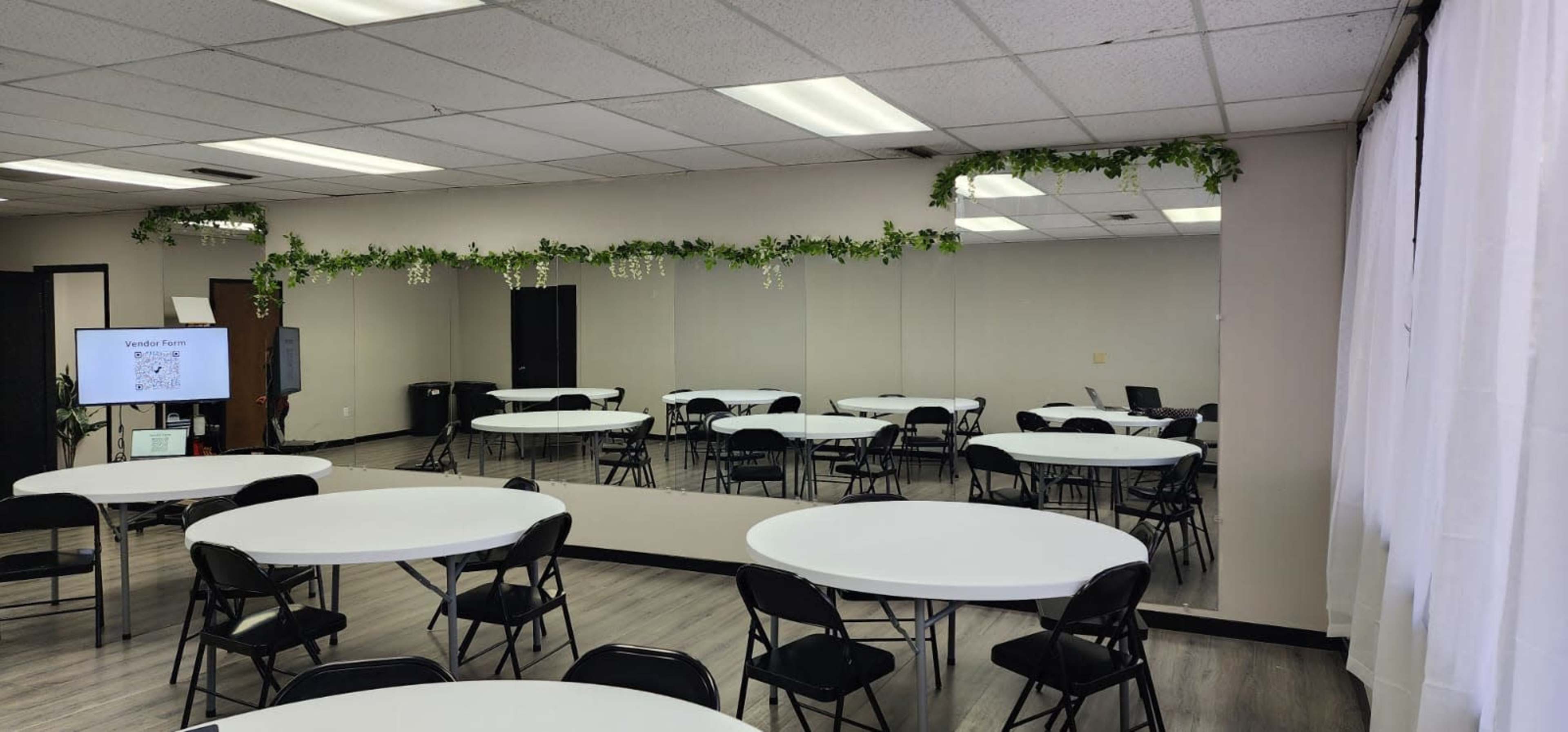 The image shows a spacious room with several round tables and black chairs arranged around them, and a long mirror on one wall with decorative greenery at the top.