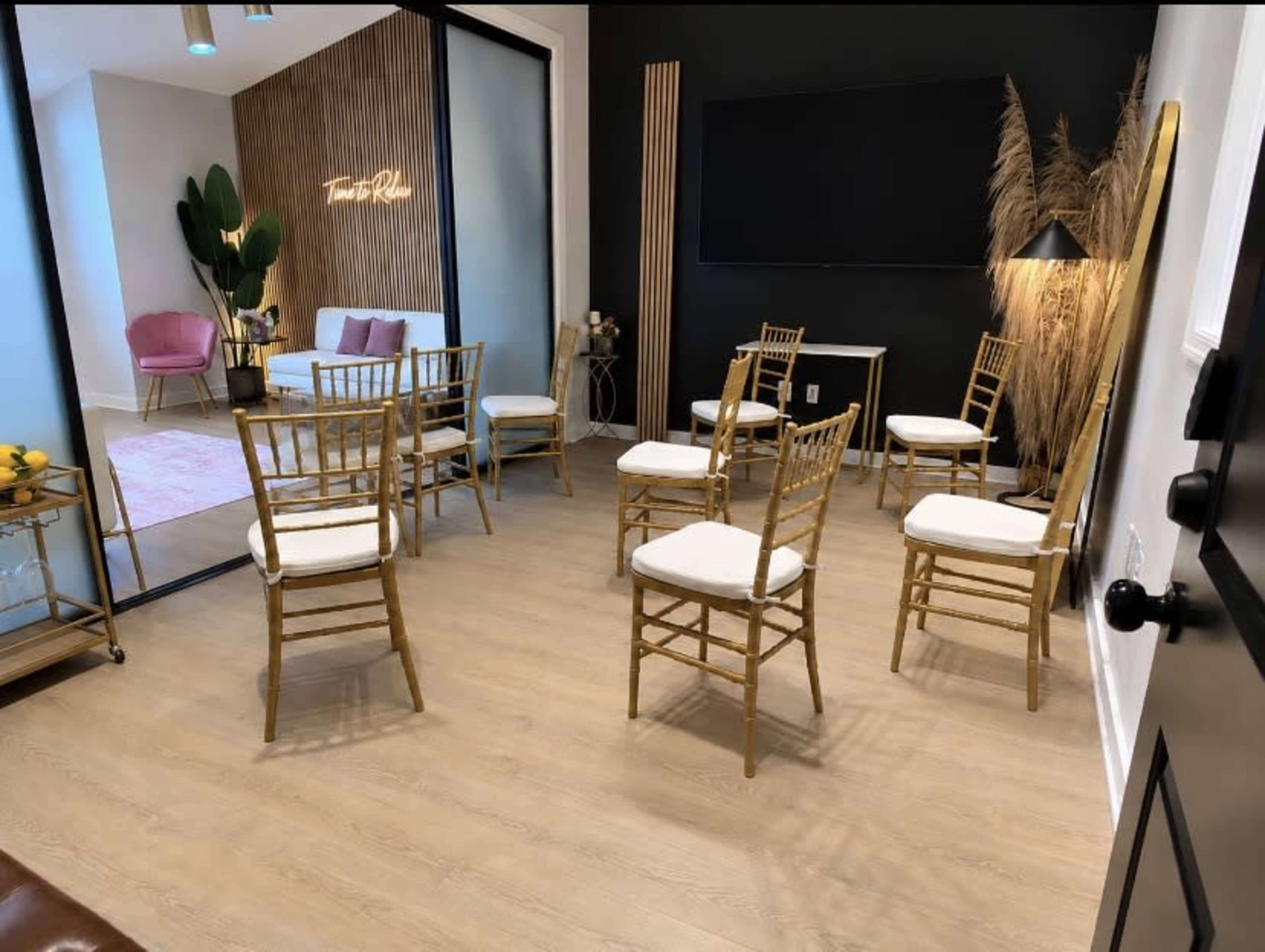 The image shows a room with golden chairs arranged in a circle on a wooden floor, with a backdrop of a dark wall featuring a television and decorative plants.