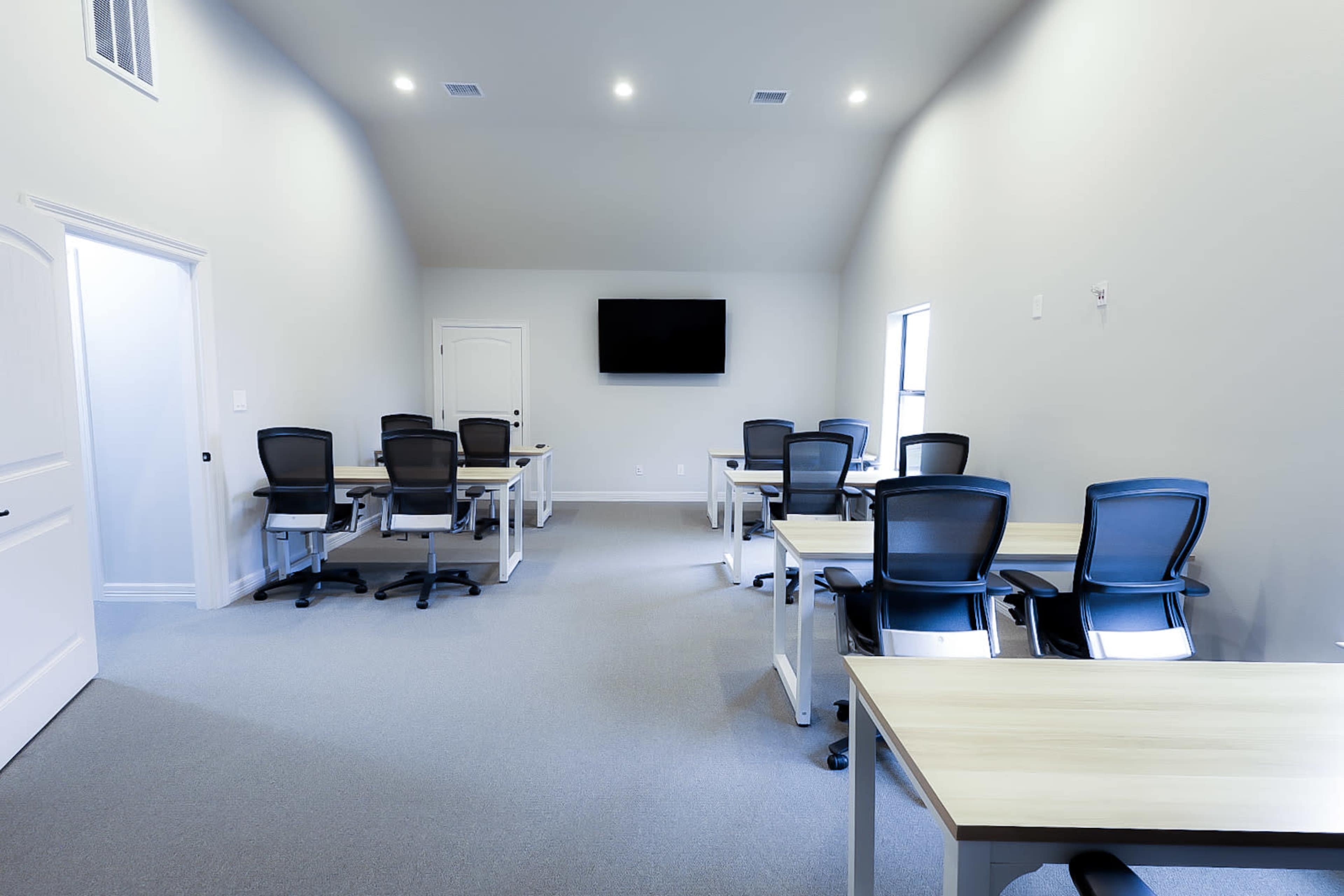 A sparsely furnished meeting room with several desks and chairs arranged in a symmetrical layout, a wall-mounted TV, and windows allowing natural light.