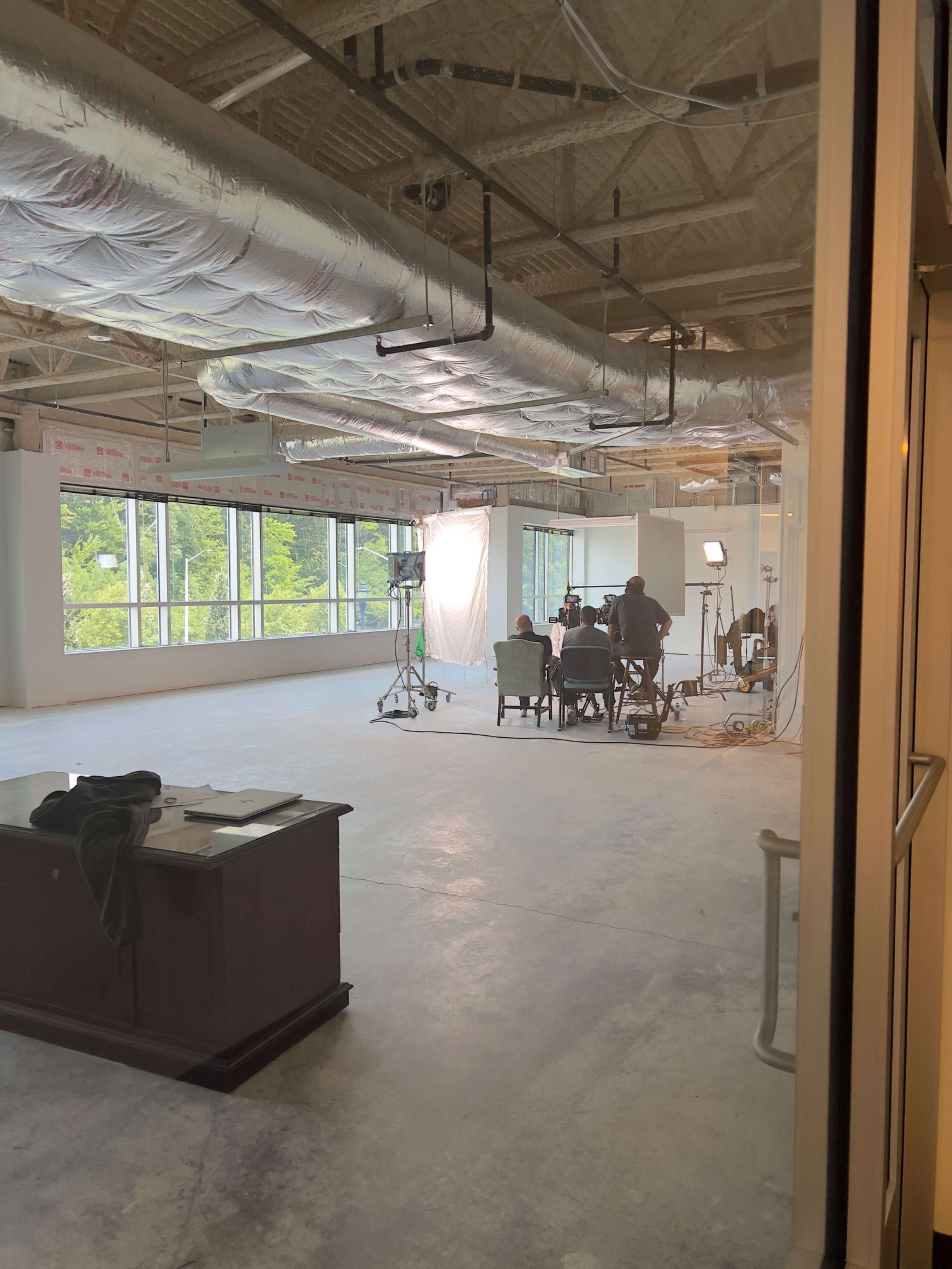 A group of people is being filmed in a spacious, unfinished office space with large windows and industrial lighting.