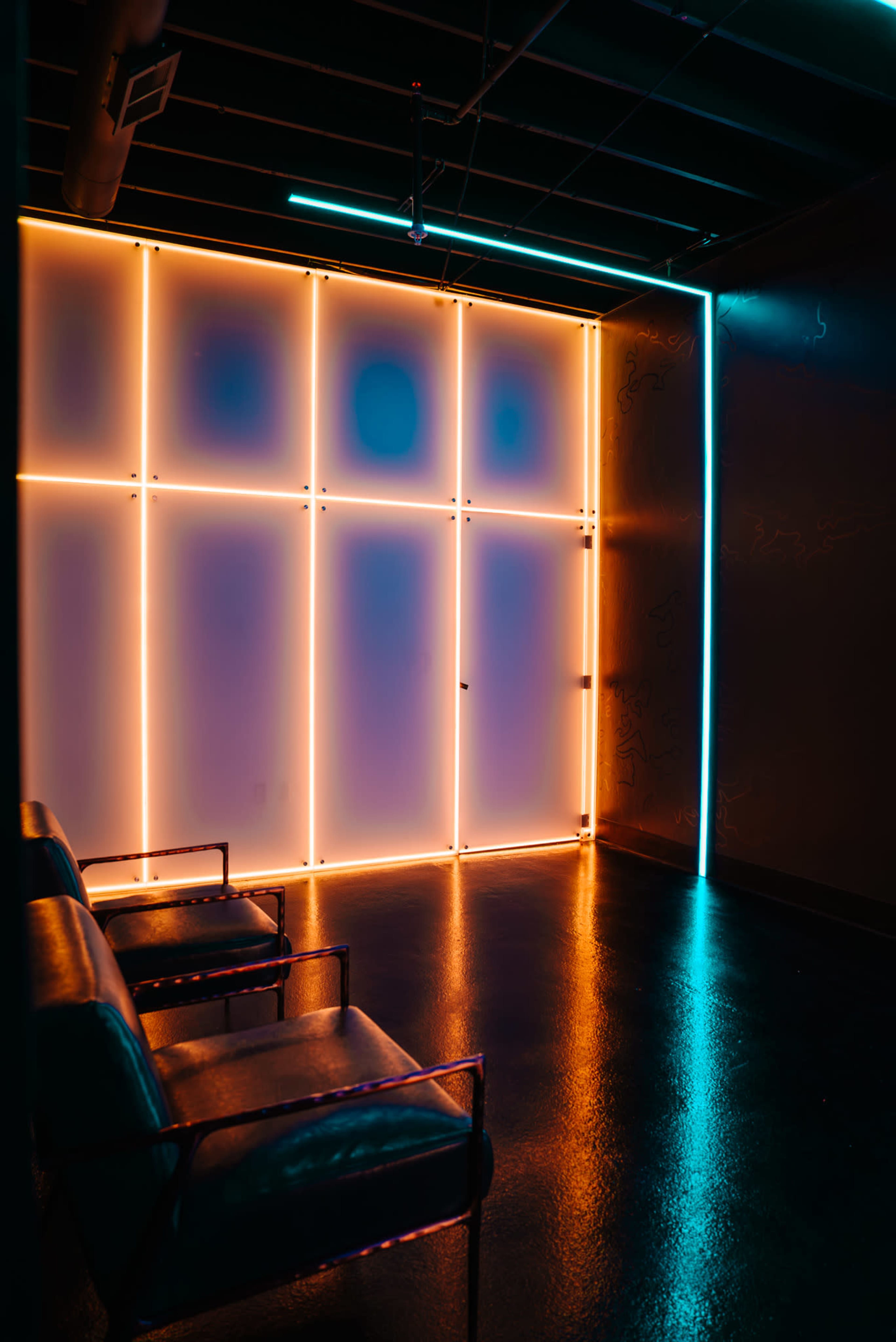The image shows a modern interior with a wall illuminated by neon lights and two black chairs placed in front of it.