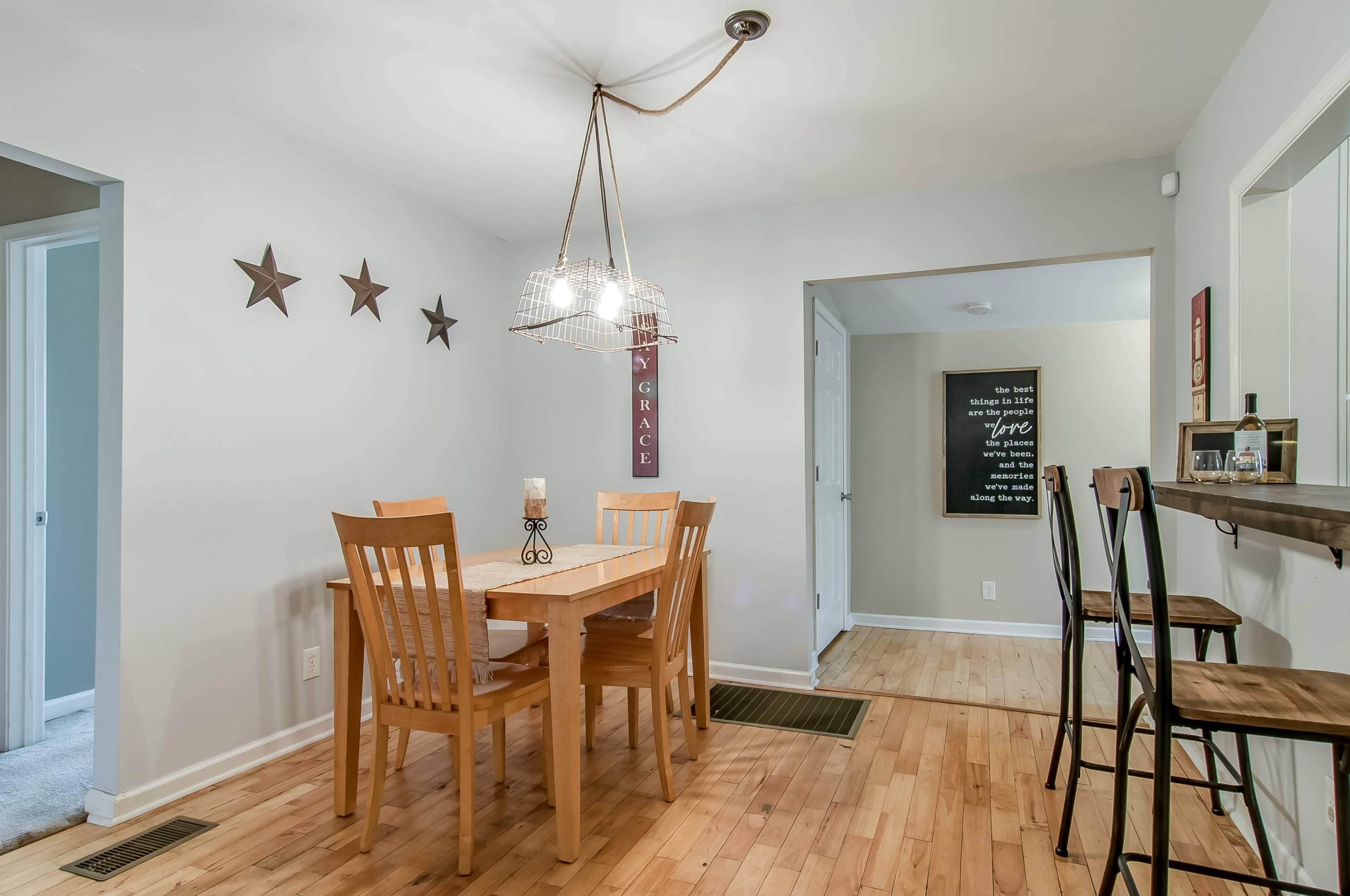 A dining area features a wooden table surrounded by four chairs, with wall decor and a small bar area in the background.