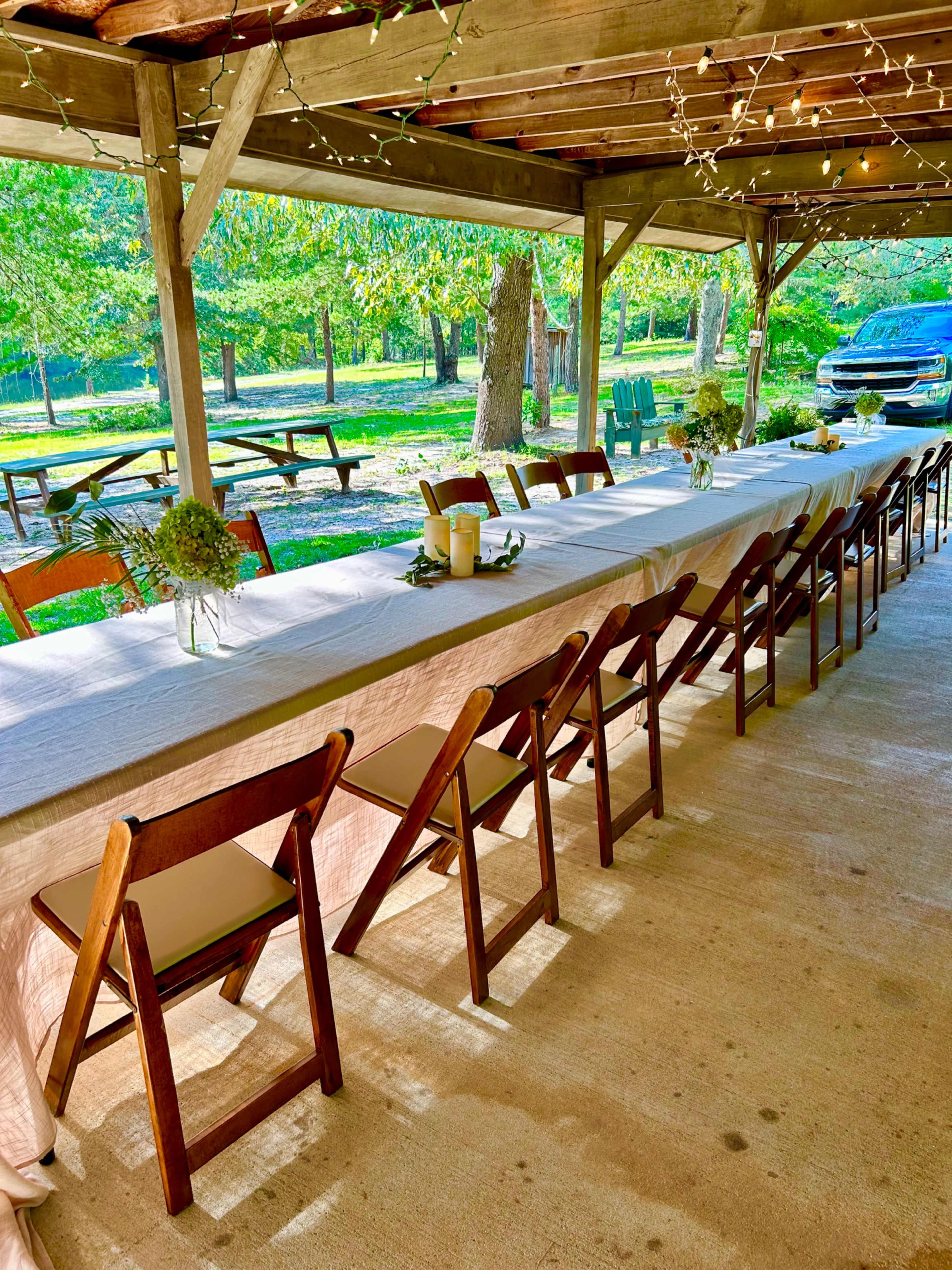 A long, covered table set up with chairs under a wooden pavilion, surrounded by trees and outdoor seating areas.