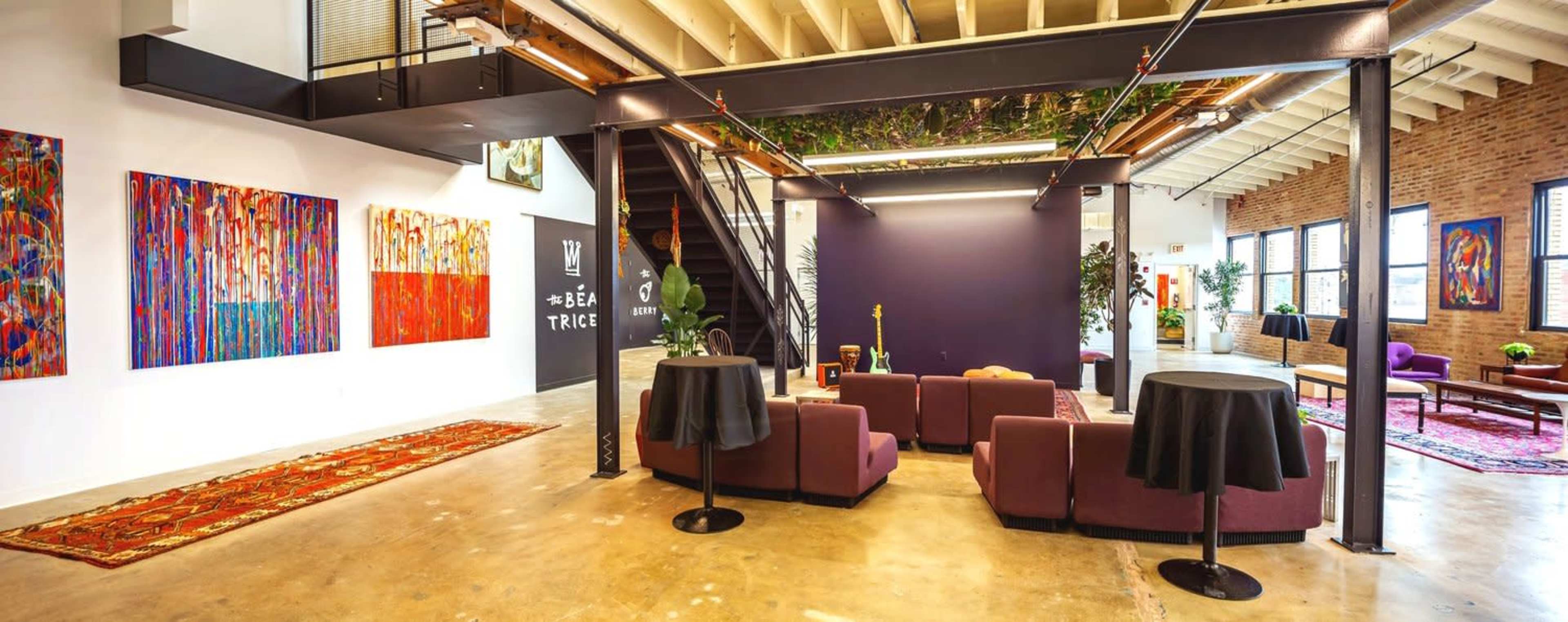 A modern art gallery with colorful paintings on the walls, seating areas featuring plush couches and tables, and a staircase leading to an upper level.