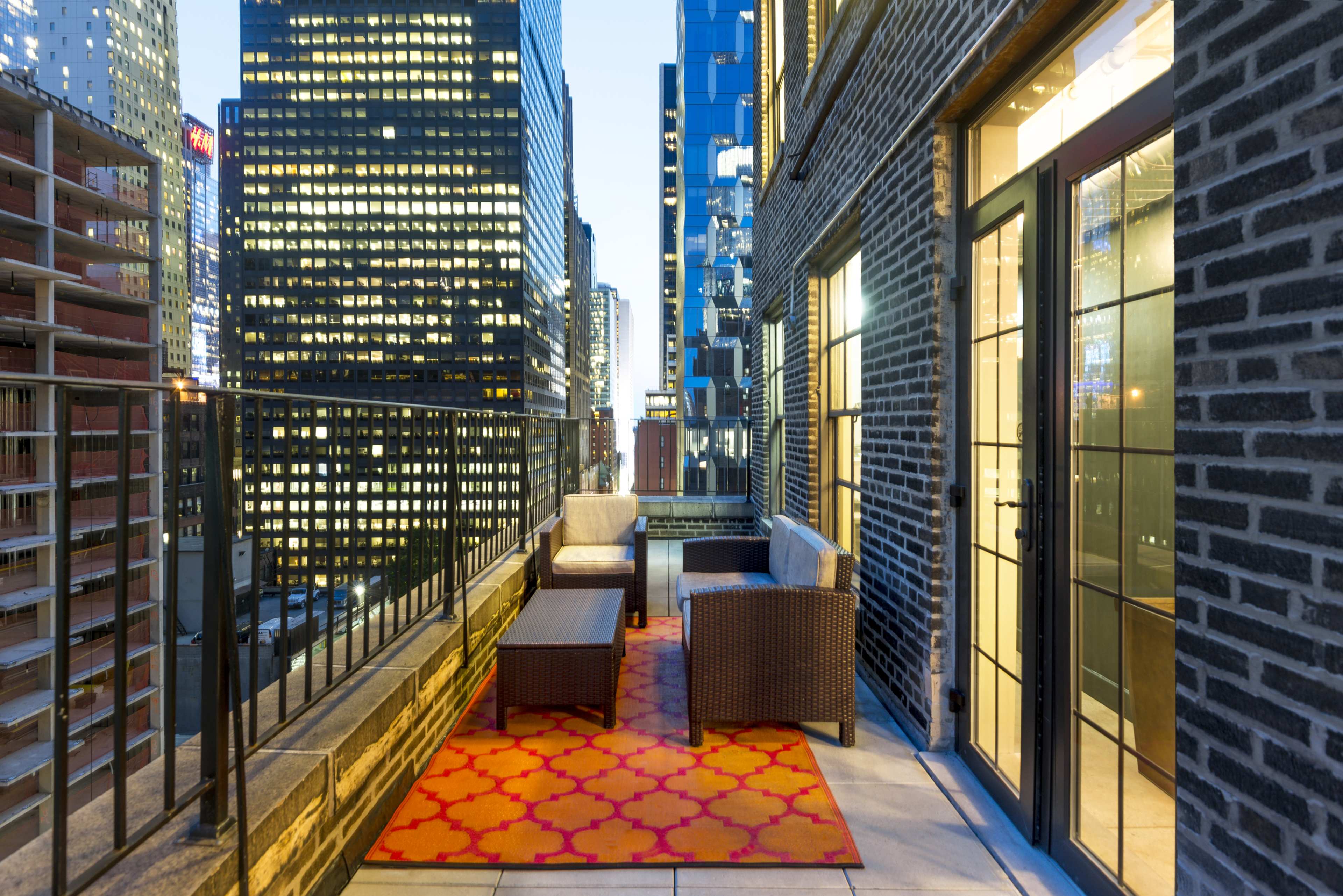 A balcony features a seating area with modern furniture, overlooking a city skyline illuminated by skyscraper lights at dusk.