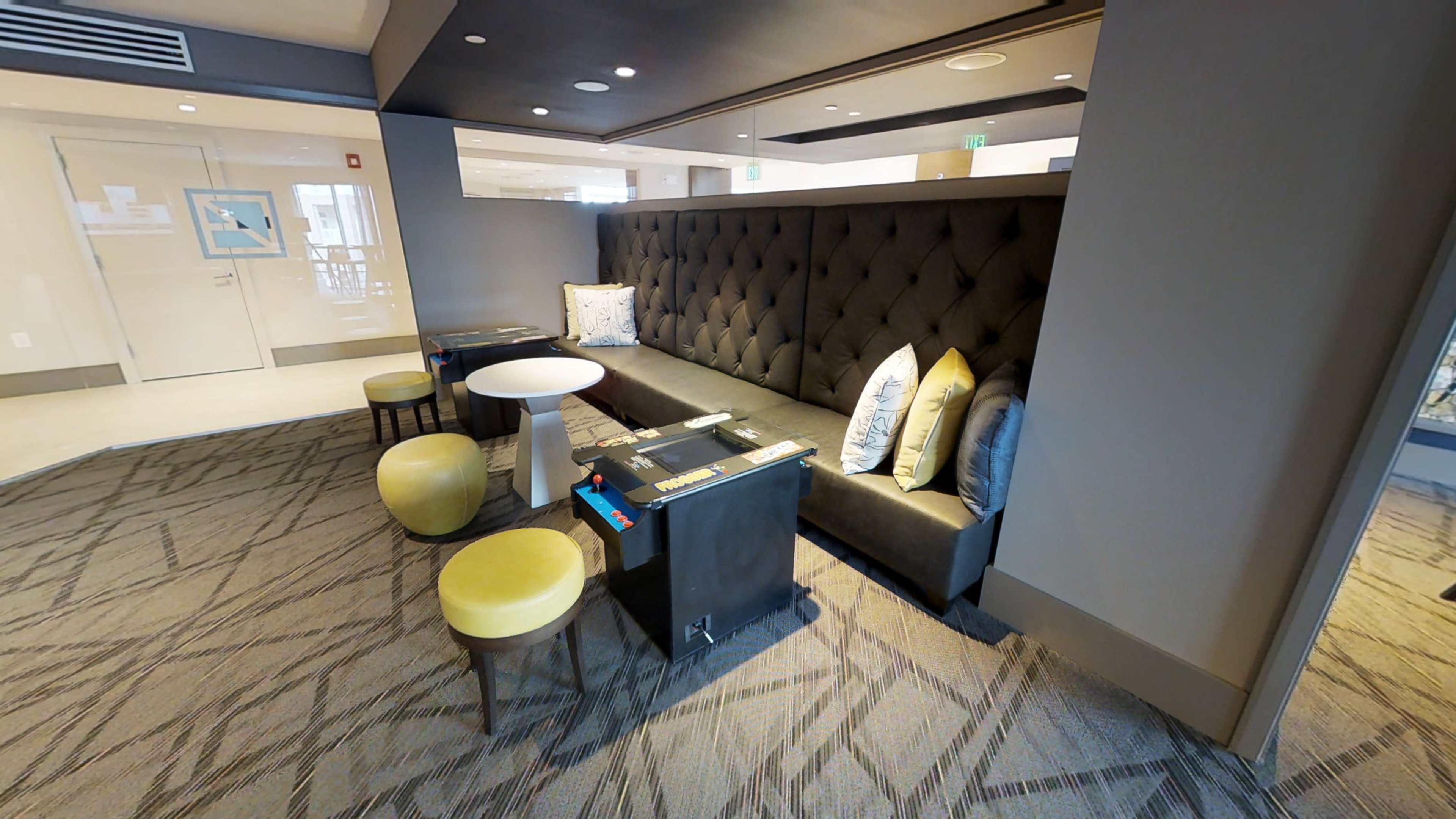 The image shows a lounge area featuring a black tufted sofa with decorative pillows, a small round table, and two round yellow stools.
