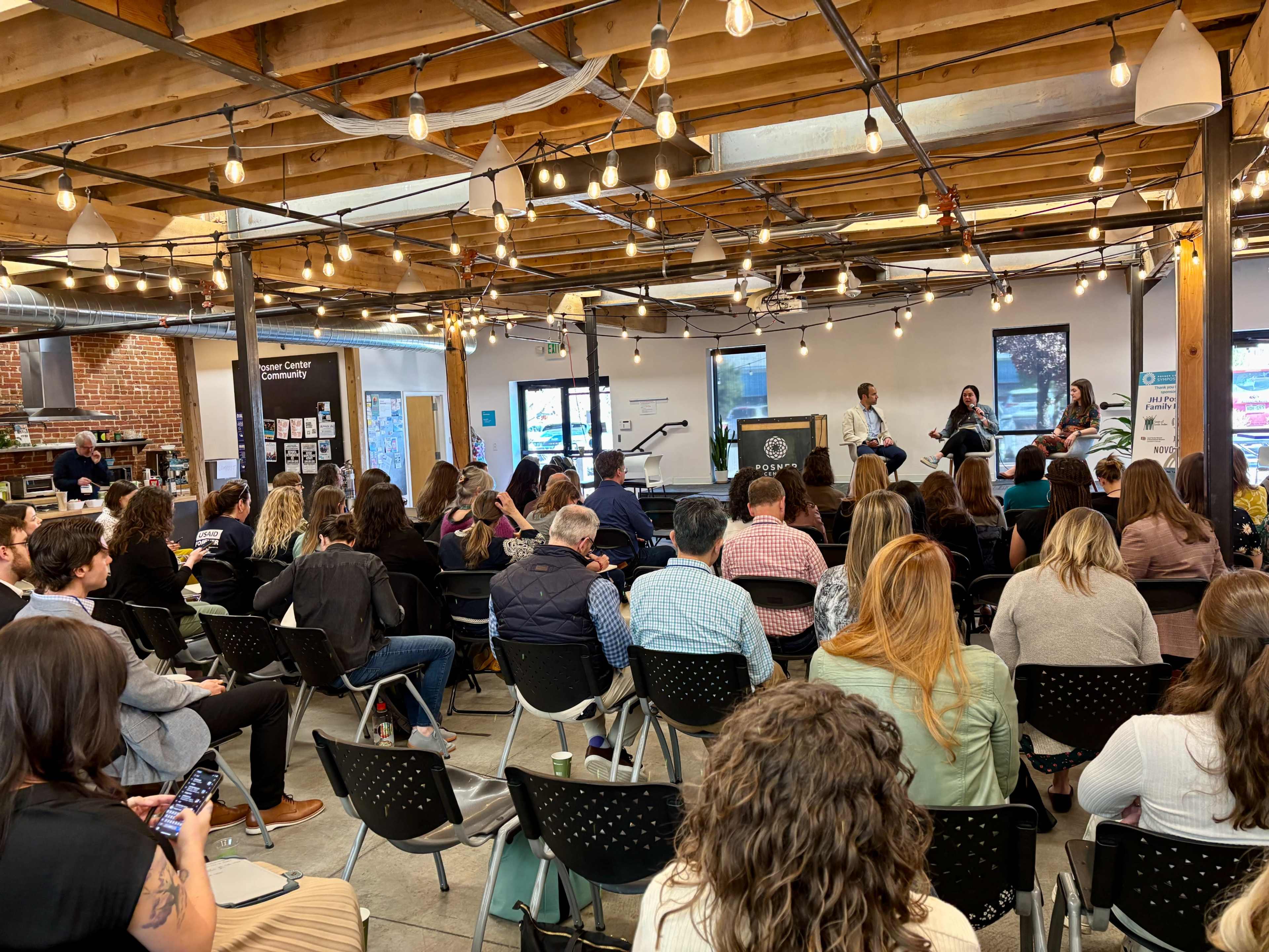 A group of people is seated in an industrial-style indoor space with exposed beams and string lights, attending a panel discussion.