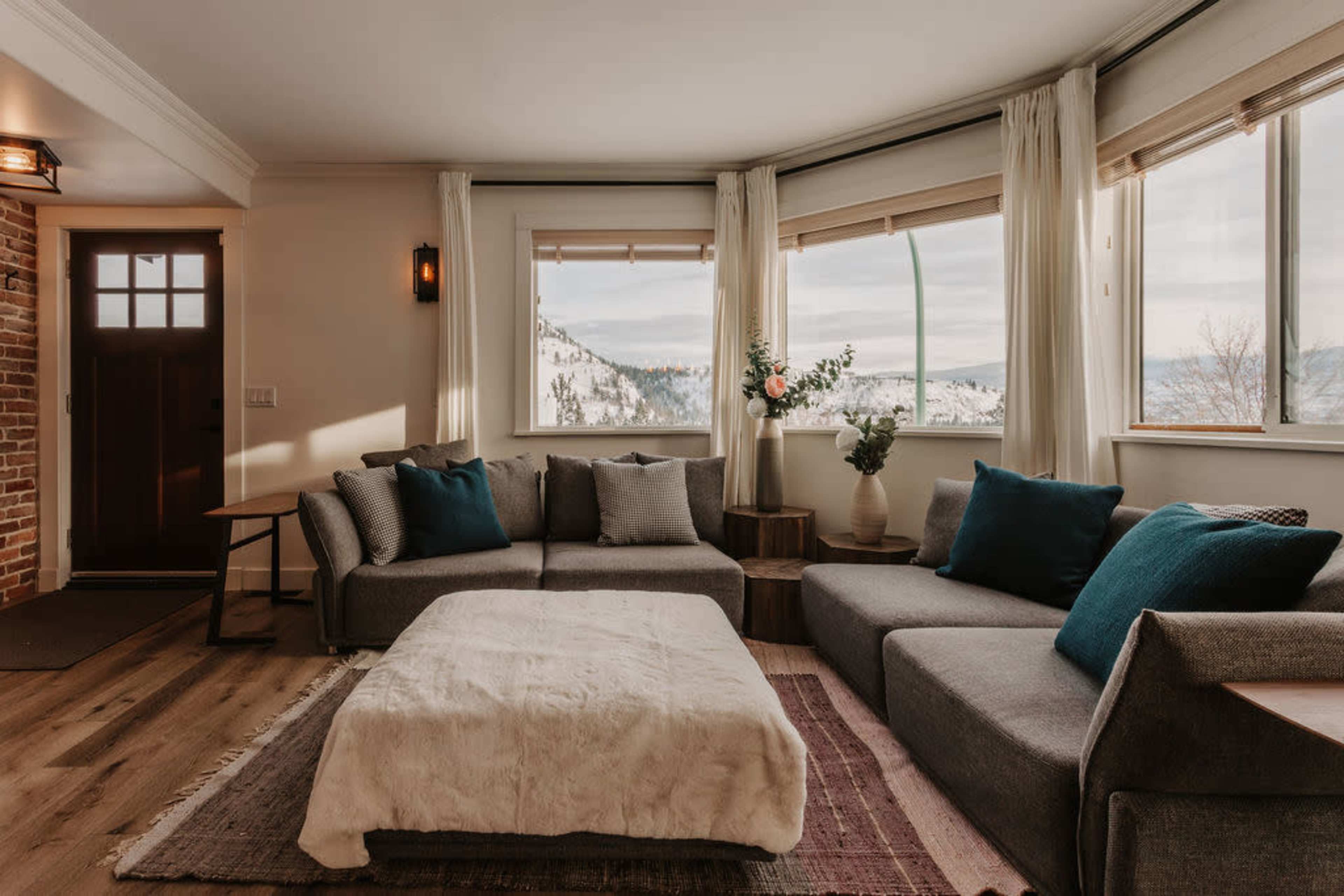 A modern living room features a gray sectional sofa with blue and gray cushions, a light-colored area rug, and large windows showcasing a snowy mountain view.