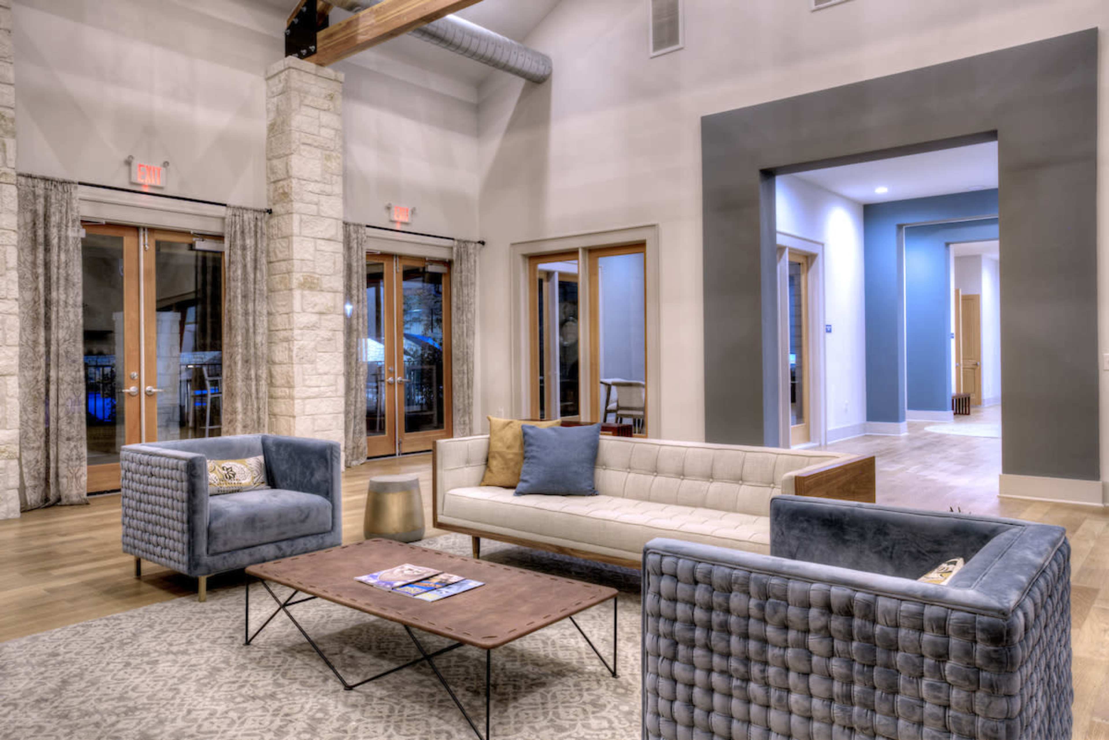 A modern living space featuring a light-colored sofa, two blue chairs, a coffee table, and large windows with curtains.