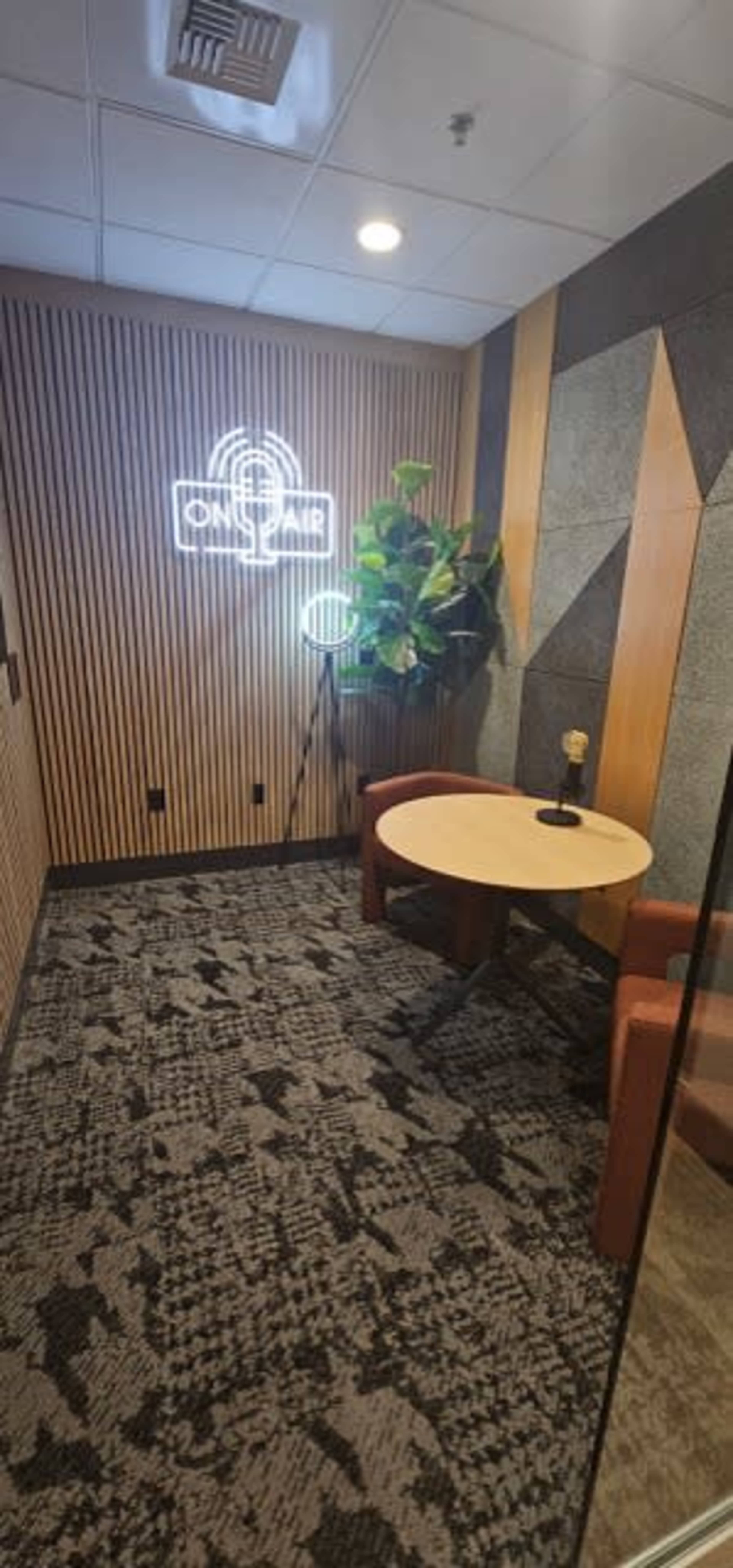 The image shows a small, modern recording studio or booth with a round table, two chairs, and a "ON AIR" neon sign on the wall, surrounded by decorative plants and wood paneling.