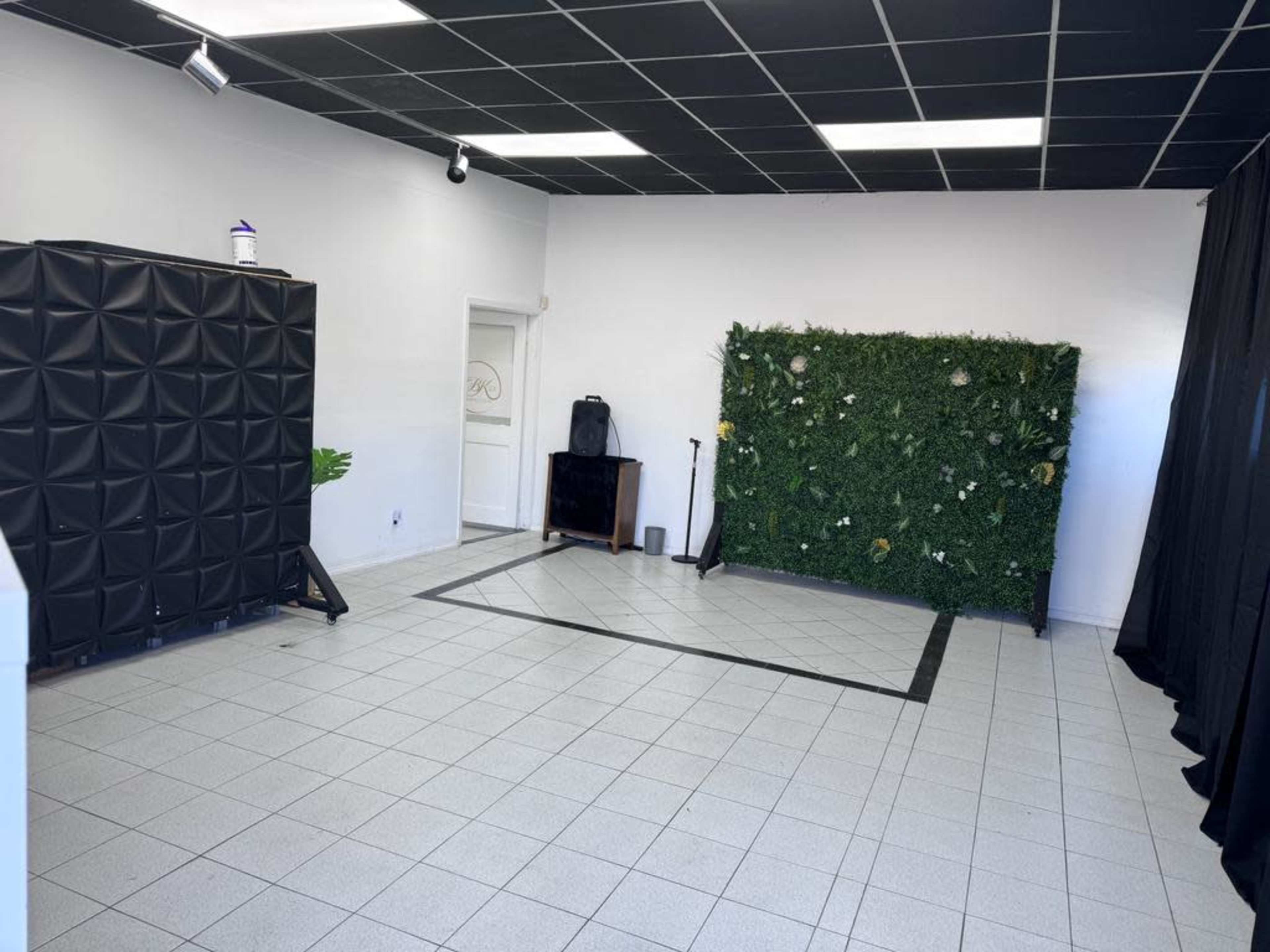 A spacious room featuring a black padded wall, a green plant wall, and a tiled floor arranged in a square layout.
