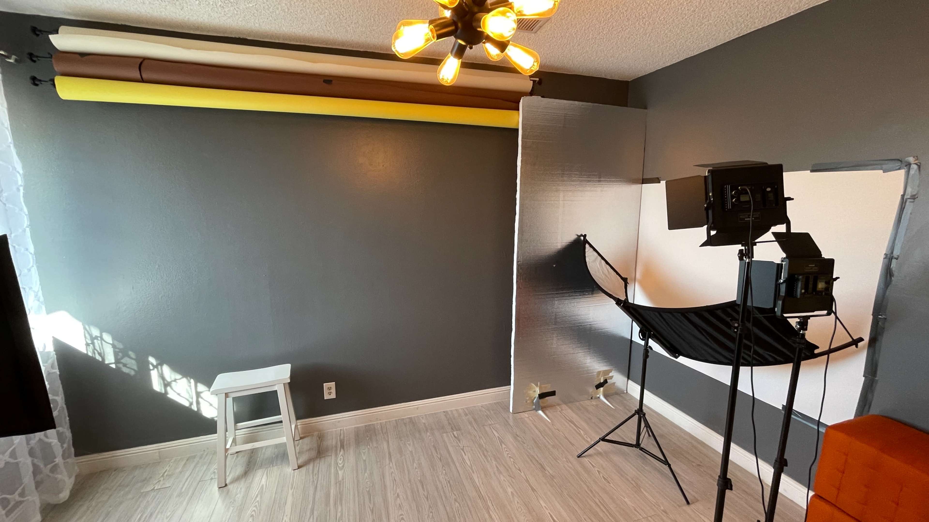 A photography studio setup featuring a stool, backdrop stands, and lighting equipment in a room with gray walls and various colored backdrop rolls.