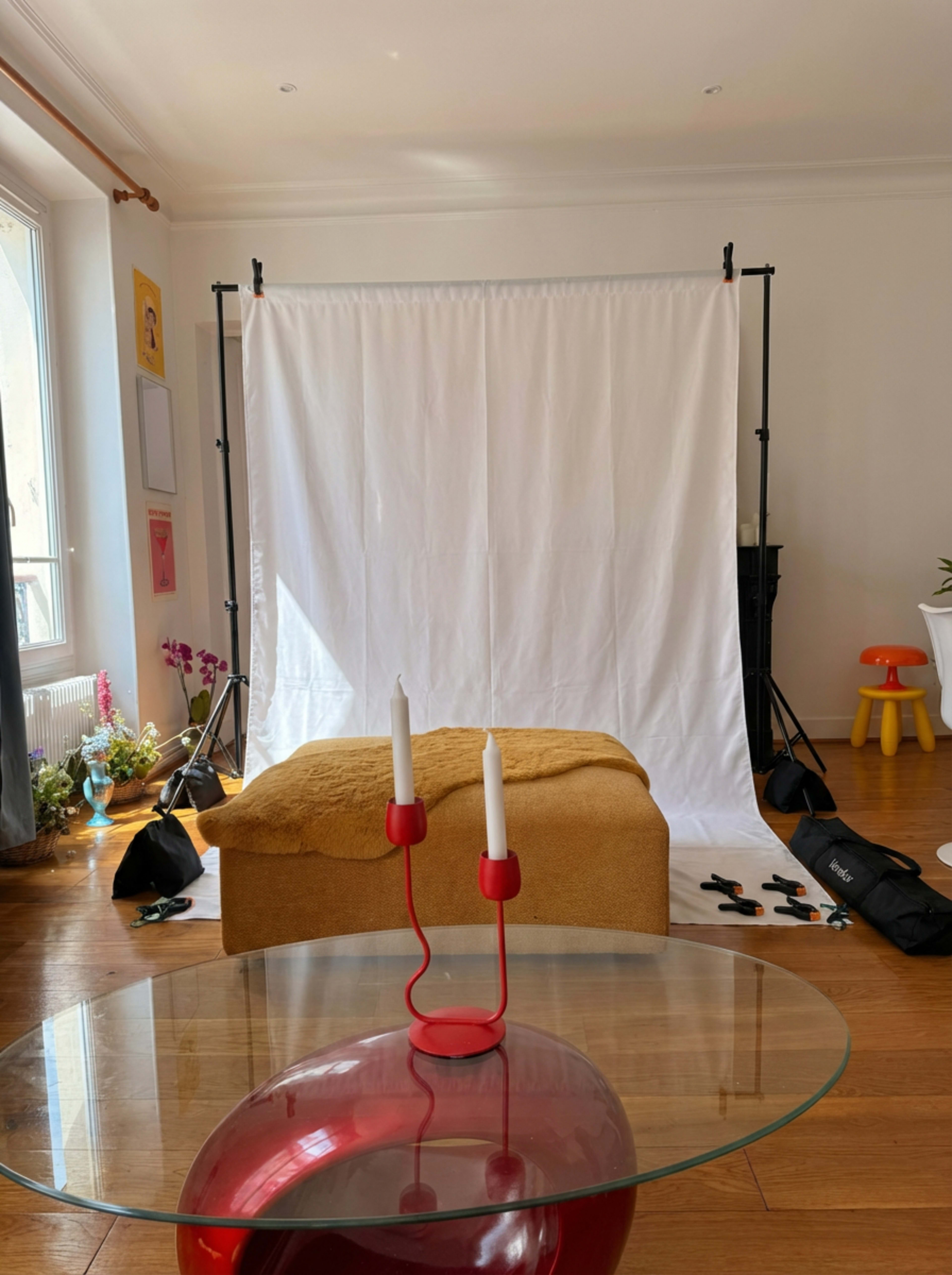 The image shows a photography setup with a white backdrop, a wooden bench covered in a yellow cloth, and a glass table in the foreground featuring a red candle holder with two white candles.