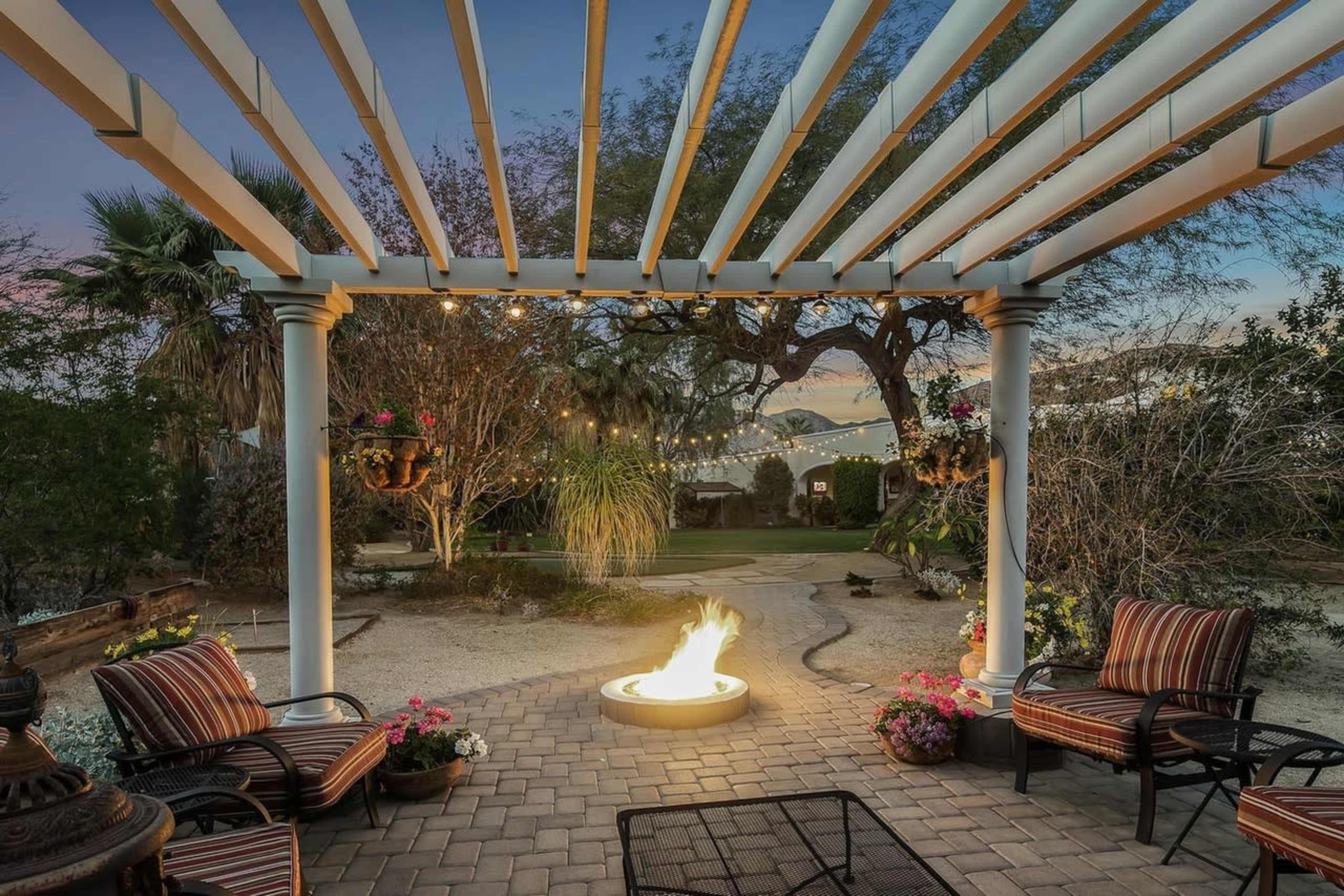 A fire pit surrounded by seating is situated under a trellis with string lights in a landscaped outdoor space.
