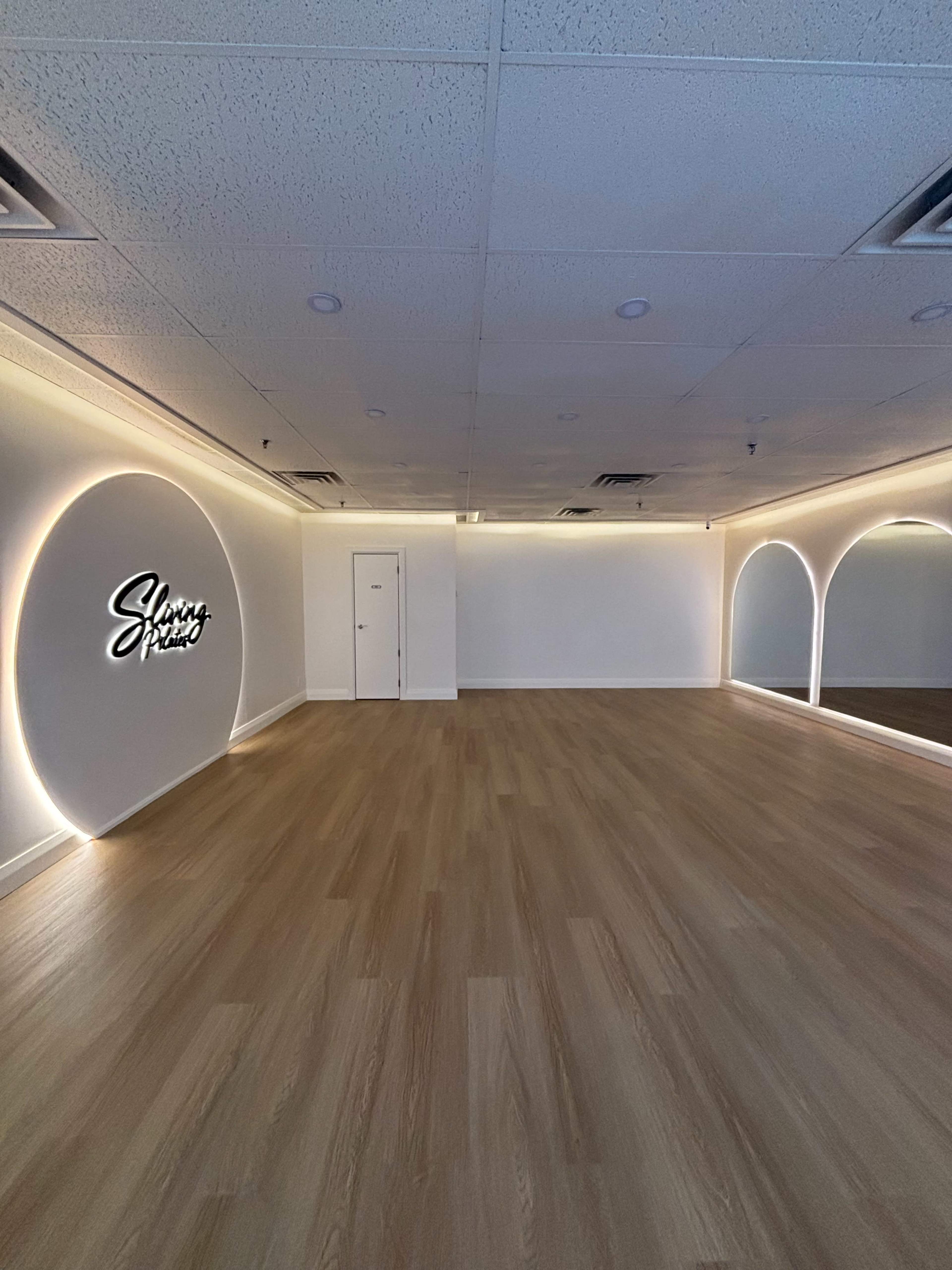 The image shows a spacious, modern room with wooden flooring, illuminated by soft light along the walls, featuring a large mirror and a sign that reads "Slimmy Peach."