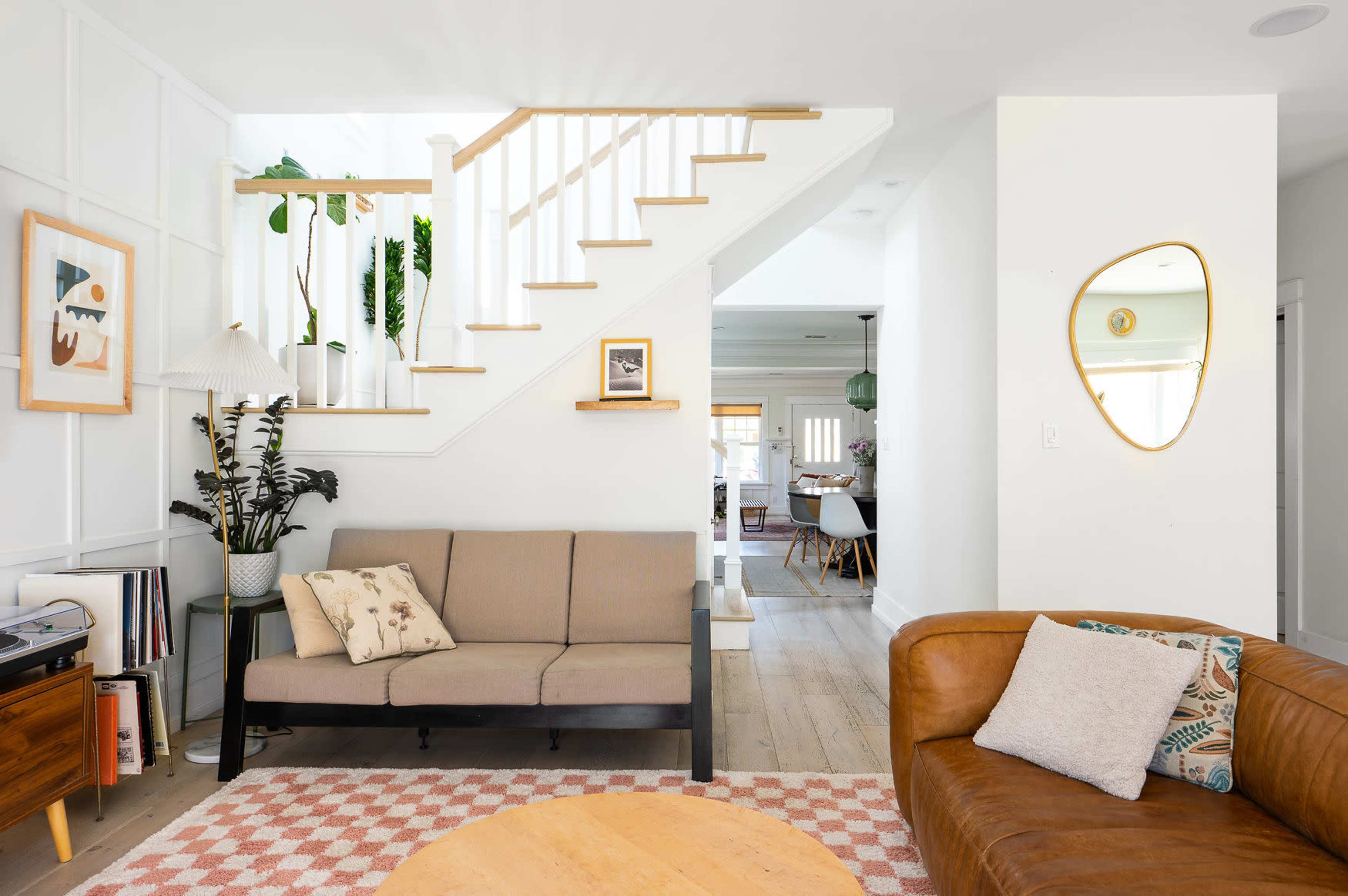 A bright living room featuring a brown couch, a beige loveseat, a round wooden coffee table, and a staircase in the background.