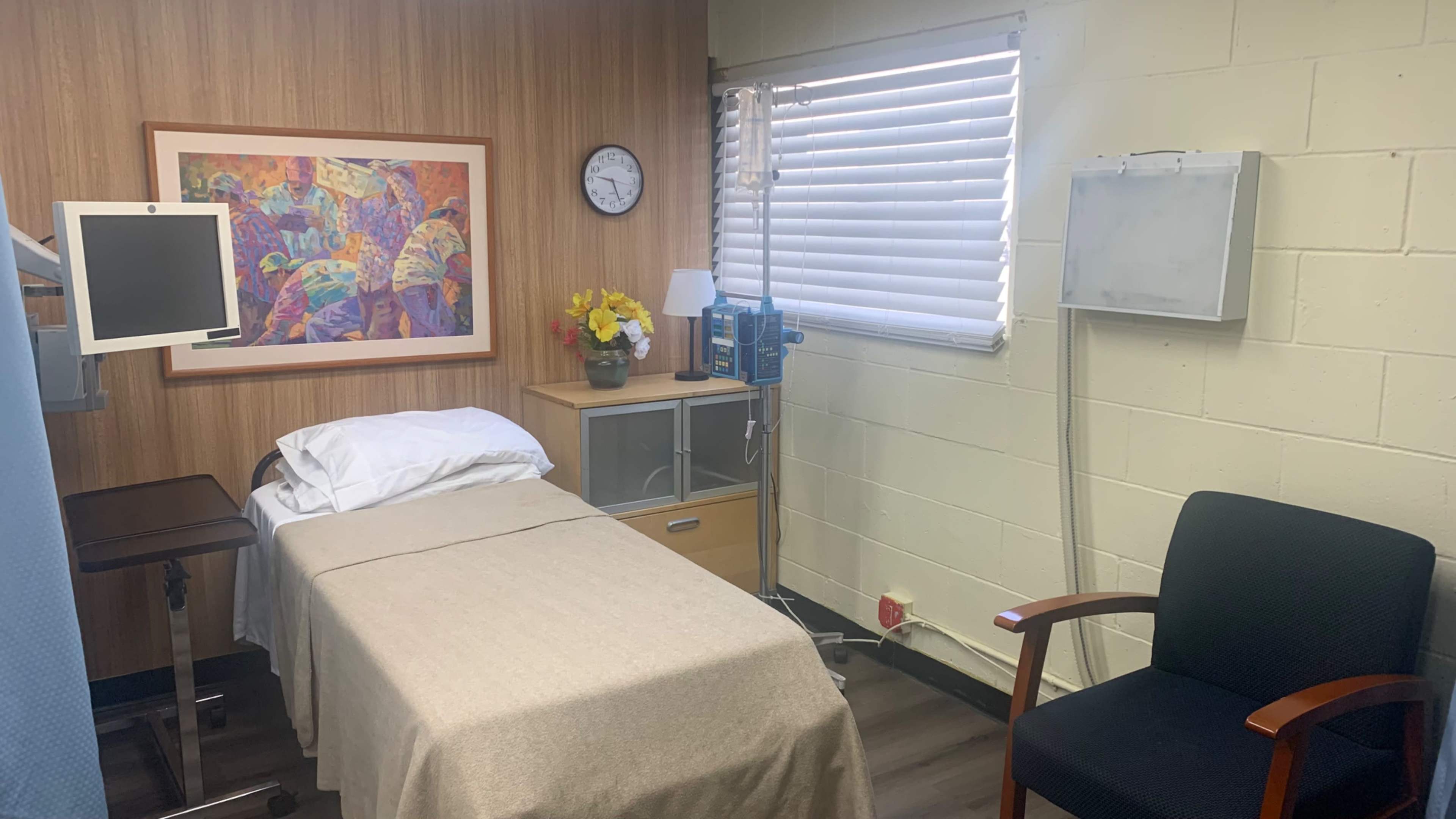 A small patient room with a bed, a chair, a wooden cabinet, and a floral artwork on the wall.