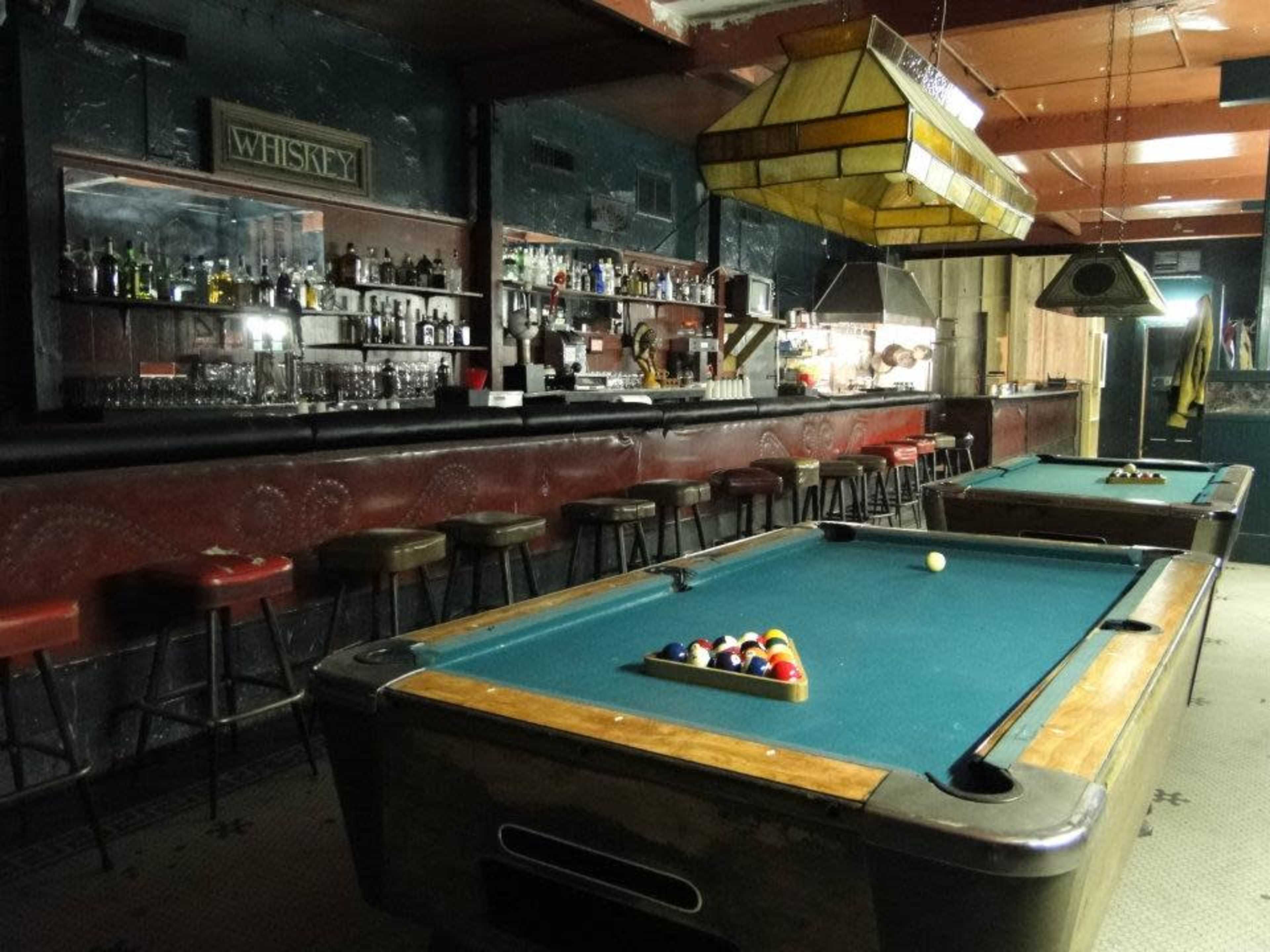 The image shows an interior of a bar with two pool tables, a wooden bar counter lined with stools, and shelves filled with bottles.